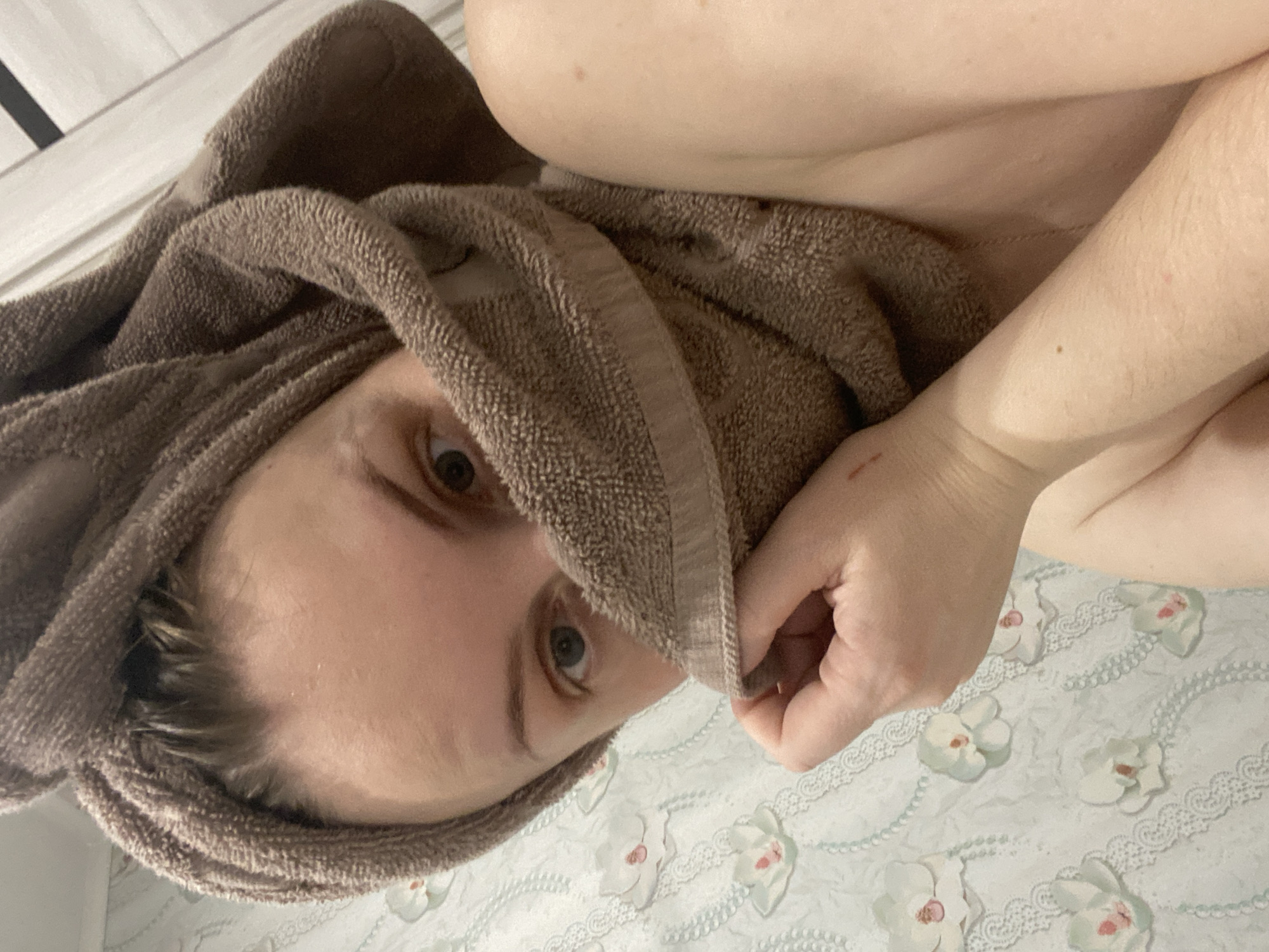 The image shows a close-up, rotated view of a person with a brown towel wrapped around their head. The person's face, particularly their eyes, is visible through the towel. Their skin appears fair. A hand is positioned near the towel. The background features a surface with a floral pattern. There is no evidence to suggest that the image was taken in the city of Noviy, Russia.