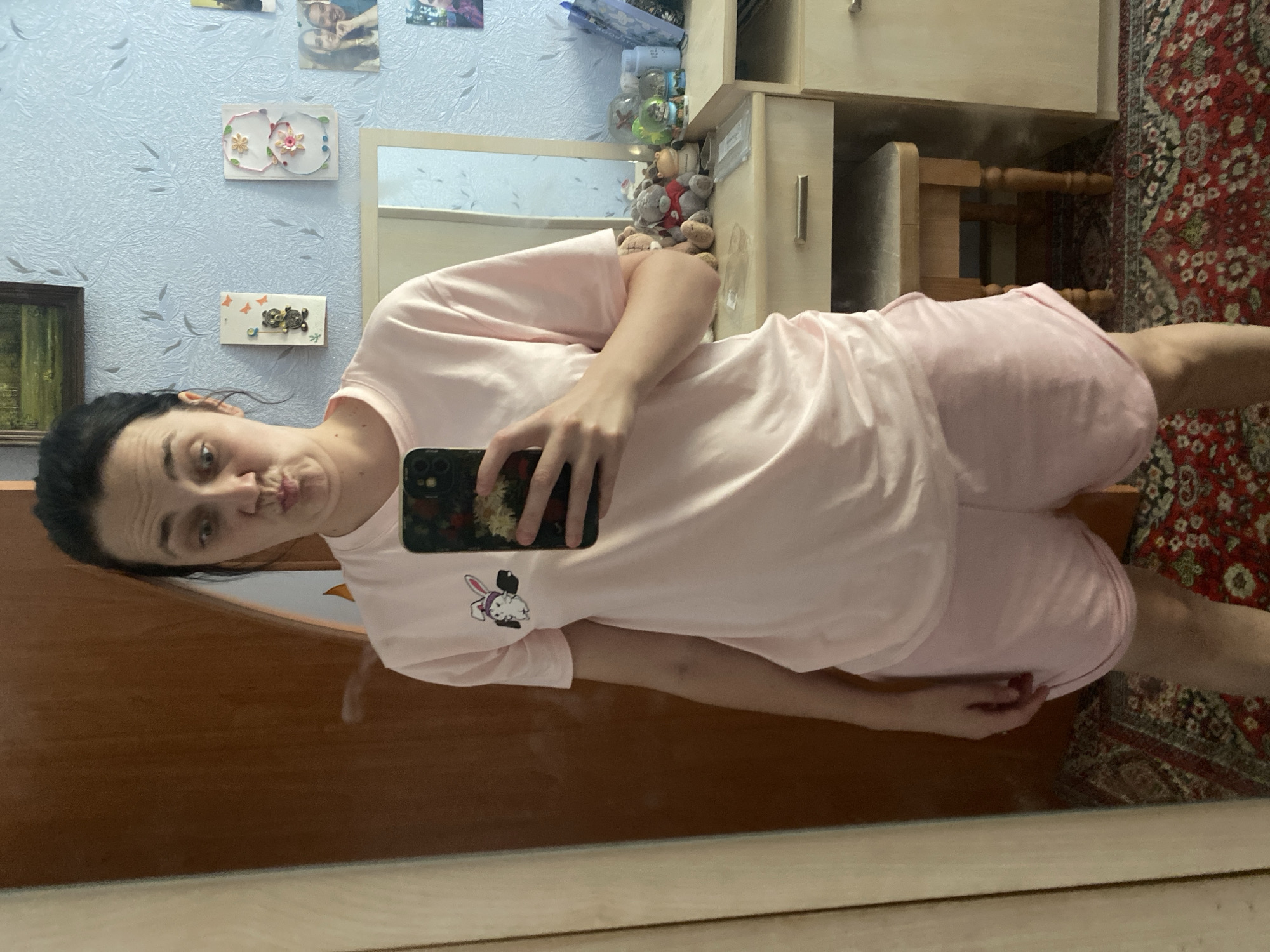 A young woman with dark hair pulled back stands in front of a mirror, taking a selfie. She is wearing a light pink t-shirt with a small cartoon bunny graphic on the left chest and matching pink shorts. Her expression is playful as she puckers her lips. The background shows a dresser with various items, including stuffed animals and decorative pictures on a blue, textured wall. A wooden door is visible to the left, and a patterned rug covers the floor. The scene appears to be in a bedroom or personal living space.