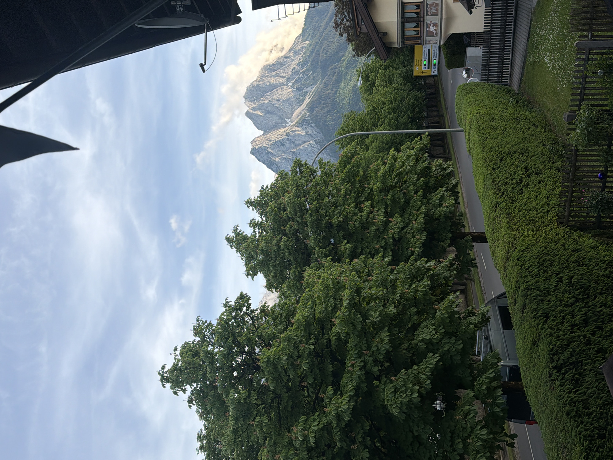 The image captures a picturesque scene of a mountainous landscape, a residential area, and a road. The perspective appears to be from an elevated position, possibly a balcony or window, looking outwards and downwards.
In the background, a large, rugged mountain range dominates, its grey, rocky peaks partially shrouded in white clouds. The sky above is bright blue with scattered, wispy clouds, indicating fair weather.
The midground features lush greenery, primarily composed of several large, mature trees with dense, dark green foliage, some showing small, light-colored blossoms. These trees line a paved road or street that runs through the scene. On the right side of the road, a neatly trimmed green hedge separates the road from a residential area. A dark wooden fence runs alongside the hedge. Further along the road, a building with traditional Central European architecture is visible, featuring ornate painted frescoes on its upper facade and multi-pane windows.
Visible objects include a black satellite dish mounted under an overhang in the upper left, alongside what appears to be a security camera or light sensor. A street lamp pole stands by the road, and a round, convex traffic mirror is mounted on a pole near the hedge. A dark-colored vehicle, possibly a van or SUV, is partially obscured by trees and can be seen on the road. Garden decorations, including blue and clear spheres, are visible in the grassy area next to the fence on the far right.
A yellow directional sign on the right side of the road lists several place names, providing a strong location cue. The visible text includes: "Garmisch-Partenkirchen", "Grainau", "Eibsee", "Seilbahn Zugspitze", "Kreuzeck", and "Alpspitz". These names pinpoint the location to the Bavarian Alps in Germany.
The scene appears to be taking place during the daytime, likely morning or afternoon, given the clear illumination. There is no discernible human activity or event occurring, portraying a serene and static landscape.