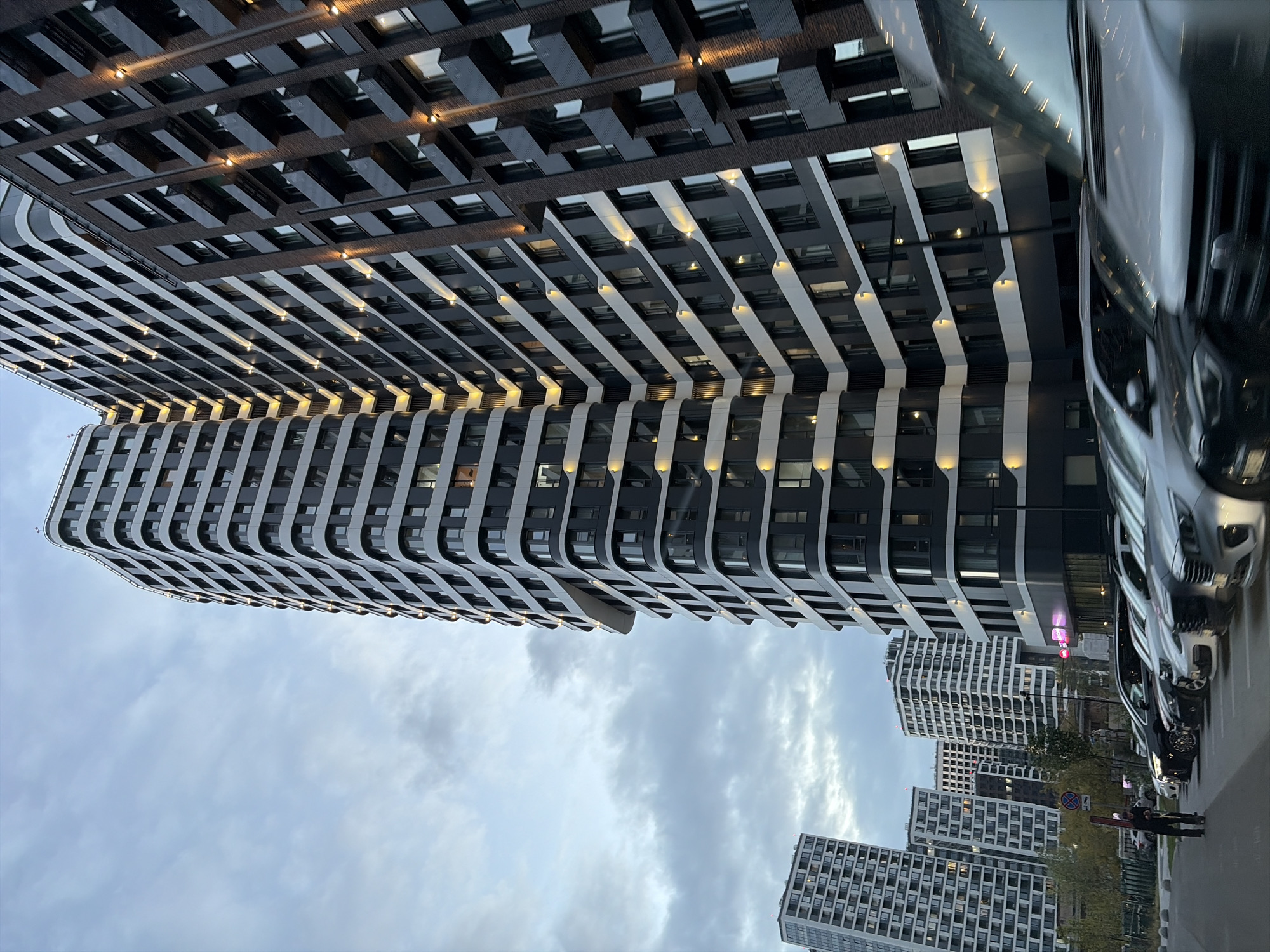 The image captures a wide-angle view of a modern residential complex in Moscow, Russia, with a cloudy sky overhead. The foreground features a row of parked cars, with a person on rollerblades visible on the left side of the frame, pulling a red object. The architecture of the buildings is characterized by their tall, multi-story structures with dark and white facades, featuring numerous windows and external lighting that highlights the building's contours. Several buildings of varying heights and architectural styles are visible, creating a dense urban landscape. Some buildings have curved facades, while others are more angular. The overall impression is one of contemporary urban development in a Russian city.
