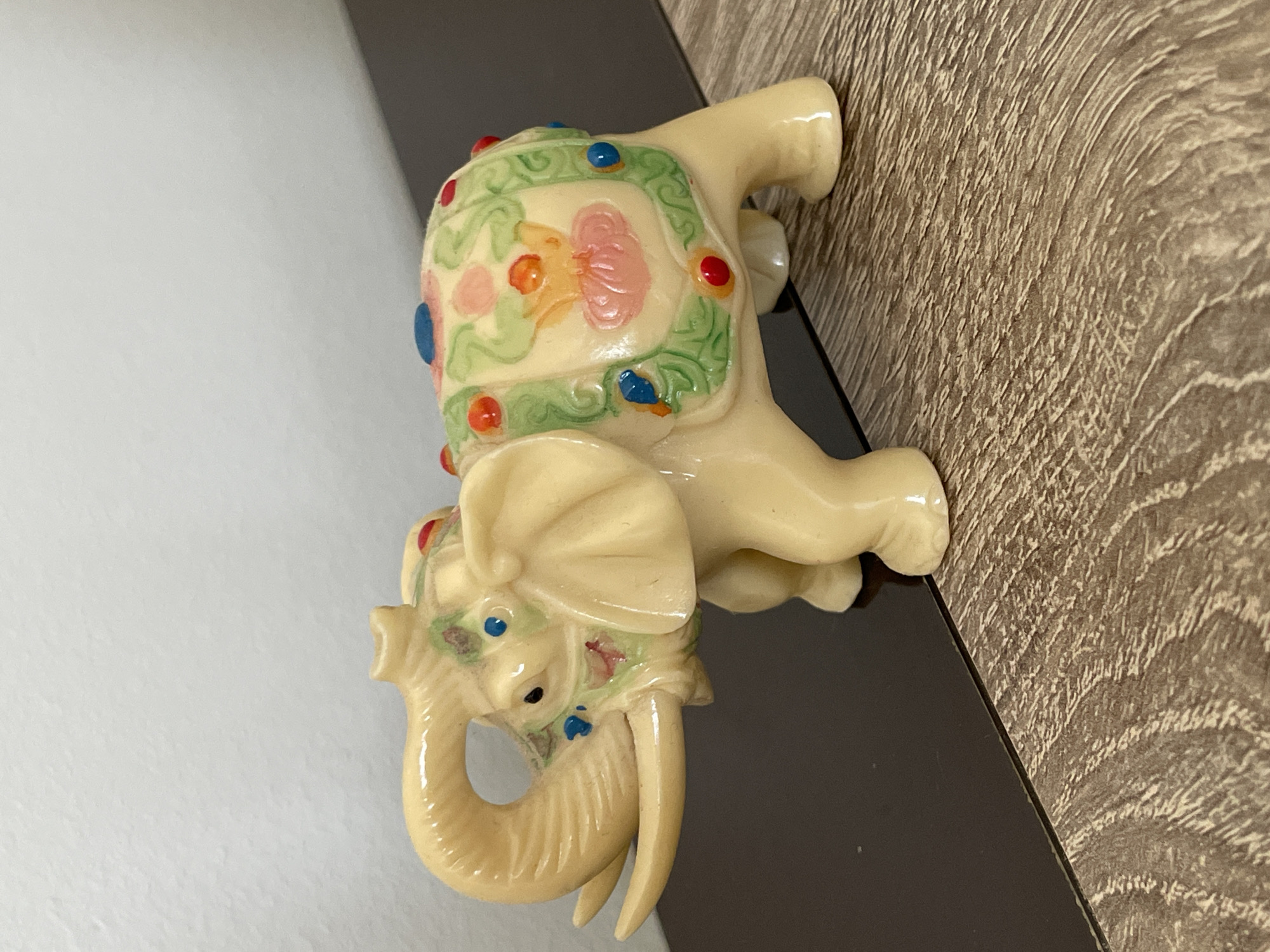 A cream-colored ceramic elephant figurine, approximately 6 inches long and 4 inches tall, is displayed on a dark brown, wood-grained surface. The elephant has a curved trunk, visible tusks, and large ears. A decorative blanket is draped over its back, adorned with painted floral patterns in green, pink, and orange, accented with small, raised red, blue, and yellow dots. The elephant is positioned facing left, with its front legs slightly forward. The background consists of a plain, light gray wall and a dark, reflective surface. The scene is indoors, likely on a shelf or table, and appears to be located in a residential setting within Trikomo, Northern Cyprus / Cyprus.