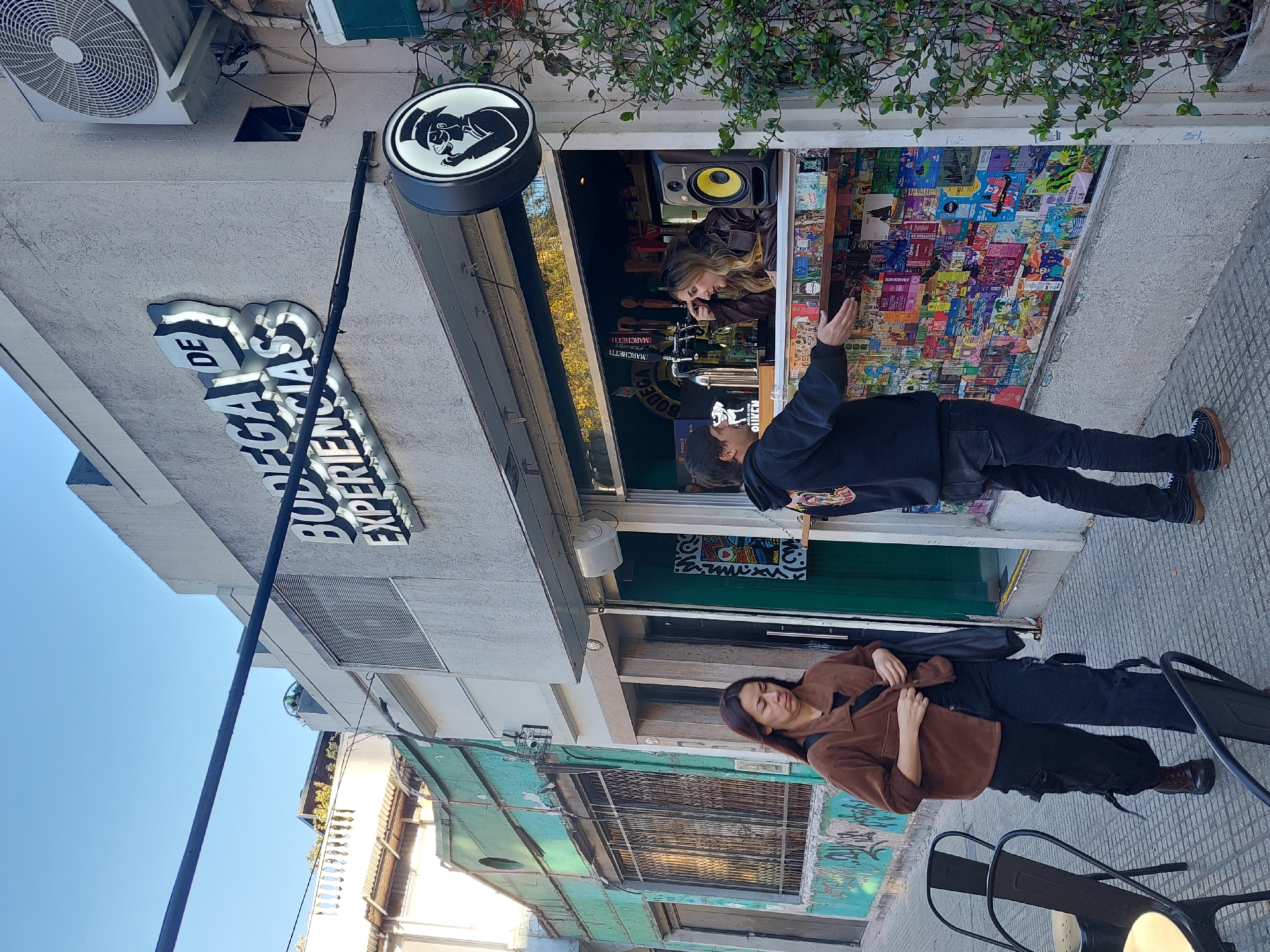 A street view in Buenos Aires, Argentina, shows a storefront labeled "BODEGA DE EXPERIENCIAS." Two people are standing in front of the store. The person near the storefront is looking up and pointing at a poster-covered wall inside the store. The other person is standing on the sidewalk. The storefront has a sign with a character on it. The buildings are a mix of concrete and painted surfaces, and the sky is blue.