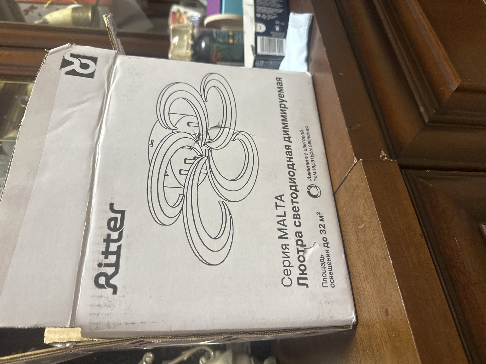 The image shows a close-up of a cardboard box with a drawing of a light fixture and text in Russian. The box appears to be for a product named "Ritter" and specifically a "Series MALTA LED dimmable chandelier." The text also indicates the area of illumination is up to 32 square meters and mentions "color temperature change of light." A logo with the letter "R" inside a square is visible in the top left corner of the box. The box is placed on a wooden surface, likely a table or furniture, with other out-of-focus objects in the background, including what appears to be packaging and some bottles. The lighting suggests it might be indoors, possibly in a home or storage area. There are no people visible in the image.