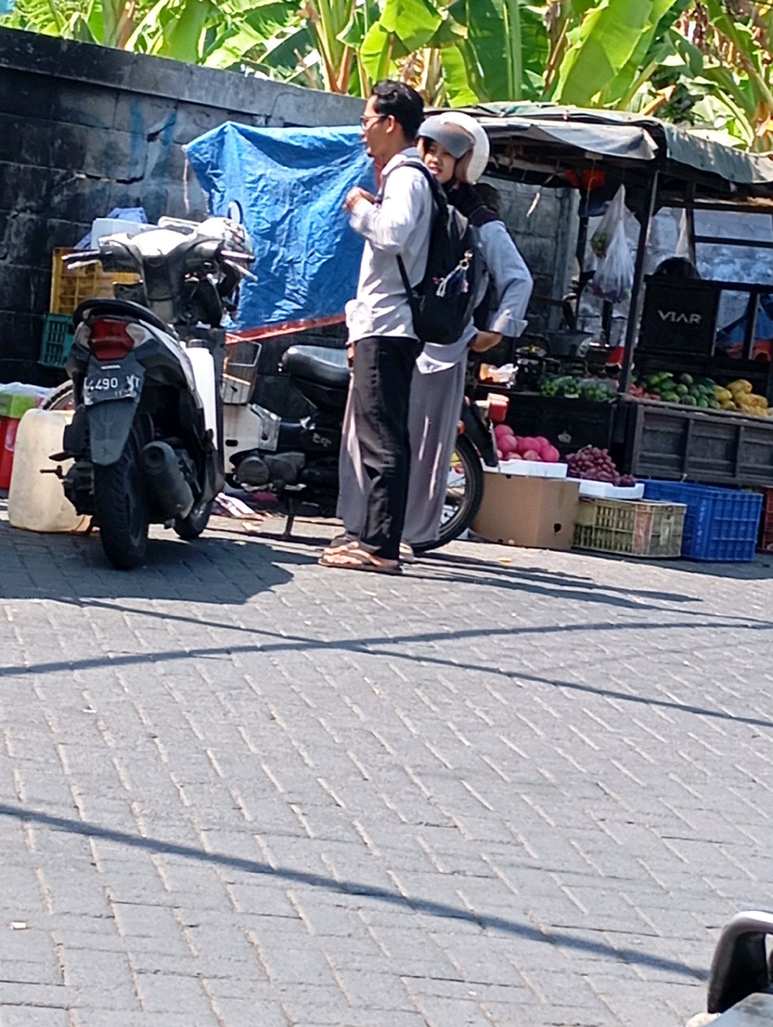 In the city of Kalisampurno, Indonesia, a man and a woman are standing beside a motorcycle on a paved street. The man, wearing glasses and a light-colored long-sleeved shirt and dark pants, is facing away from the camera and looking towards the right. The woman, wearing a light blue long-sleeved shirt and a helmet, is facing the man and has her arm around his shoulder. A backpack is slung over the man's left shoulder.
Behind them, a market stall is set up with various fruits and vegetables on display. Some of the produce includes red grapes, pink-colored fruit that could be rambutan or lychee, yellow fruits that might be mangoes or papayas, and green fruits that resemble apples or pears. The stall has a dark-colored wooden structure and is covered by a light-colored tarp. Several blue and green plastic crates are visible in front of the stall.
A white and black scooter is parked to the left of the people. The license plate of the scooter reads "L 4490 YT". To the left of the scooter, a large white container is situated. The background features a grey, rough-textured wall with a blue tarp draped over it. Lush green foliage, possibly banana leaves, is visible above the wall, suggesting a tropical environment. The ground is paved with interlocking grey bricks. The scene appears to be in daylight, with shadows cast on the ground.