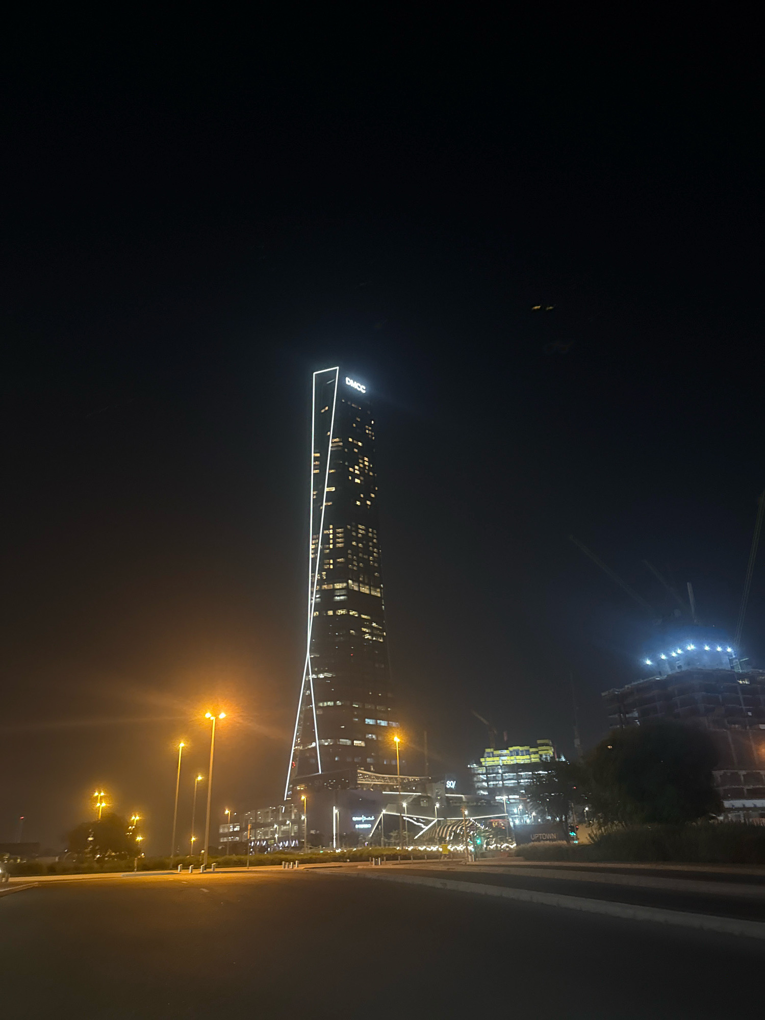 The image captures a nighttime view of the DMCC skyscraper in Jumeirah Lakes Towers, Dubai, United Arab Emirates. The iconic tower, illuminated with white lights that trace its angular design, stands tall against a dark, hazy sky. Multiple floors of the skyscraper are lit, indicating activity within. In the foreground, a street is visible, bordered by streetlights casting an orange glow. To the right of the skyscraper, construction sites are apparent, with cranes and partially built structures, including one with a bright, halo-like lighting. The overall atmosphere is urban and modern, with the city lights creating a dramatic nocturnal scene.