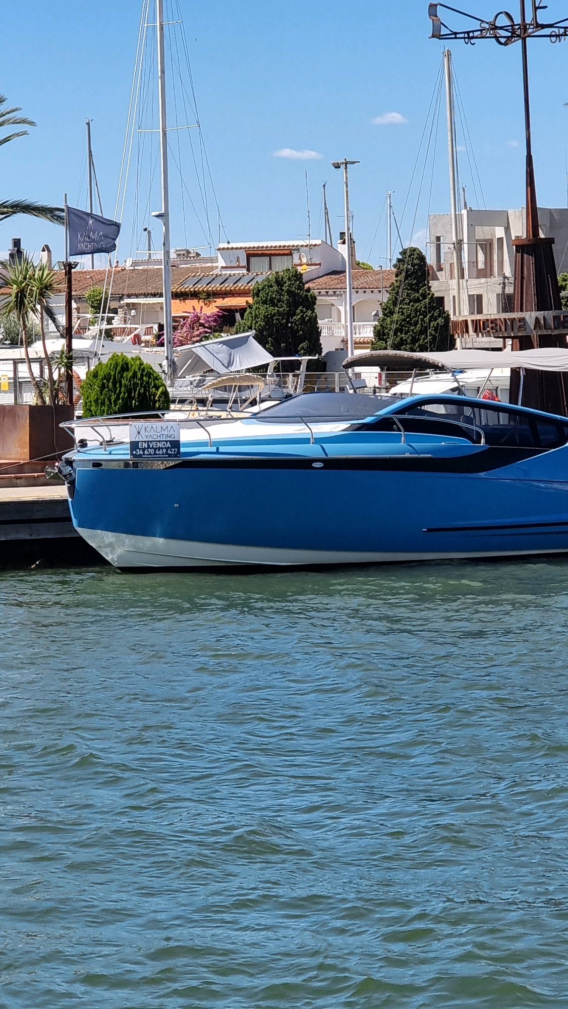 The image depicts a blue and white motorboat docked on a body of water, likely a canal or marina, in Castelló d'Empúries, Spain. The boat has a sleek, modern design with a dark-tinted windshield. A sign with the logo "KALMA YACHTING" and contact information, including a phone number "+34 670 469 427", is affixed to the side of the boat. Behind the boat, several other vessels, including sailboats with tall masts, are visible. The background shows buildings with terracotta roofs and palm trees, characteristic of the Mediterranean climate. The sky is clear and blue with a few wispy clouds. The overall scene suggests a sunny day in a tranquil, upscale marina.
