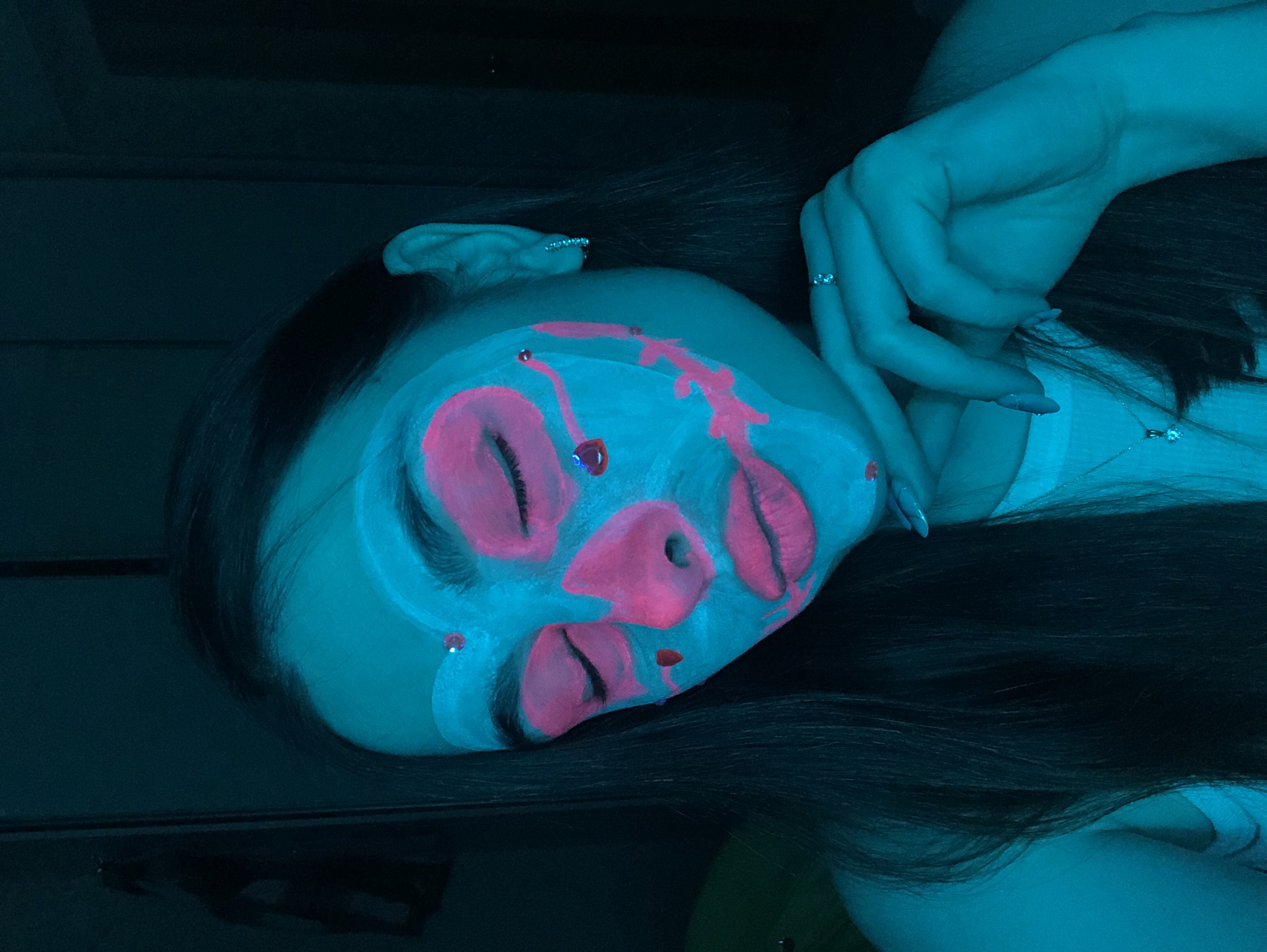 An upward-angled, close-up shot captures a person with elaborate, glowing face paint under ultraviolet (UV) light, likely in Noviy, Russia. The individual, appearing to be female, has long, dark hair that frames the face and cascades over the shoulder. Her face is predominantly painted in a vibrant, glowing blue-green hue, with striking neon pink accents that highlight features like the eyes, lips, and nose in an abstract, artistic manner. Pink "drips" or stylized patterns extend from the forehead and cheeks, and small, shimmering gems or glitter adorn the face, catching the light.
The person's eyes are closed, adding a serene or contemplative quality to the pose. Her left hand is visible on the right side of the frame, resting gently with fingers subtly curled. Several rings with small, reflective stones are visible on her fingers, and her fingernails are painted a light color. A delicate necklace with a small, sparkling pendant is visible near her collarbone, partially obscured by her hair.
The overall lighting is dark and moody, characteristic of a blacklight environment, which causes the specific makeup pigments to fluoresce vividly. The background is indistinct and dark, emphasizing the subject. The image suggests an artistic photoshoot, a costume event, or a party where UV-reactive makeup is used. There is no visible text in the image.