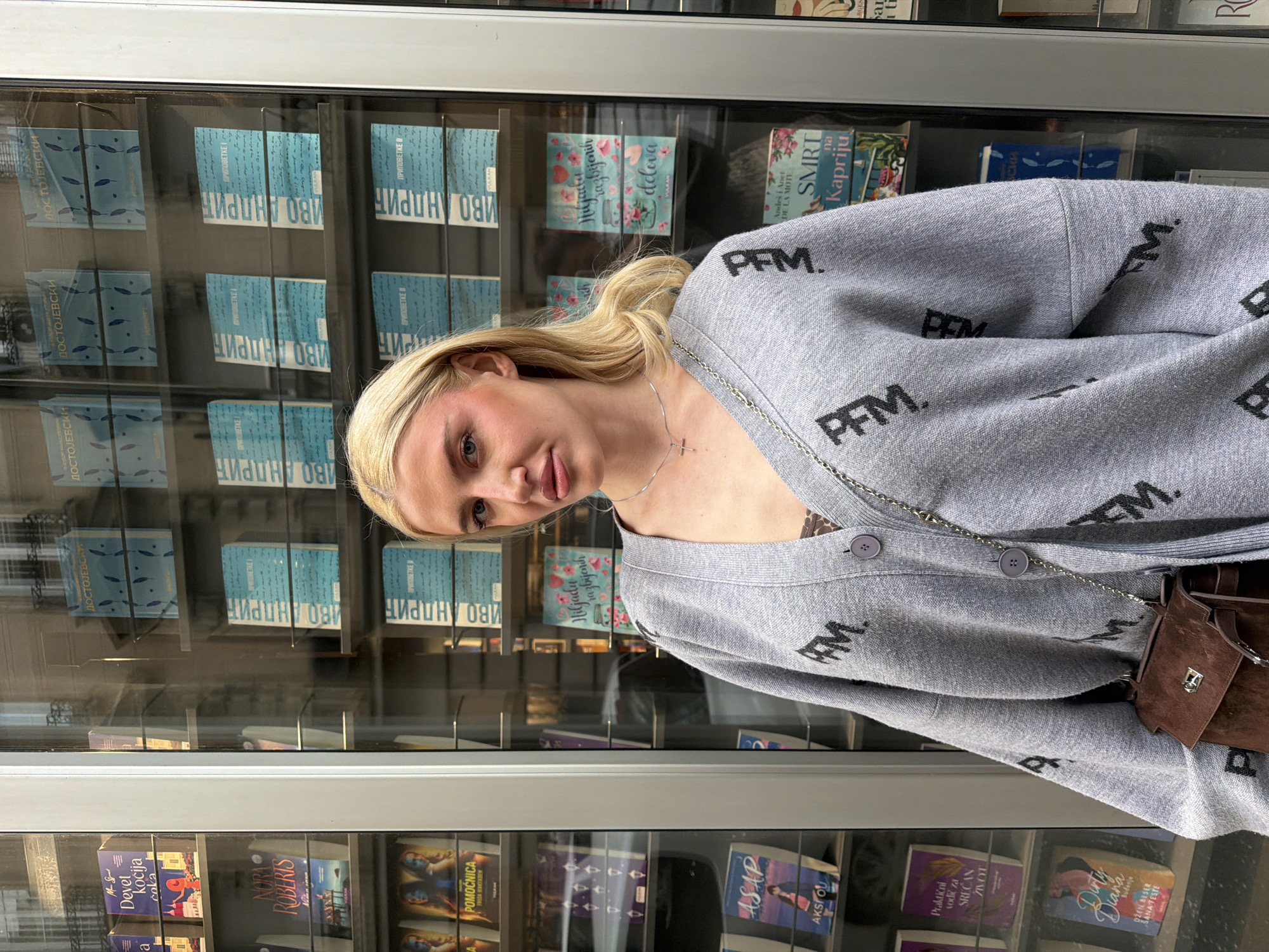 A young woman with blonde hair and a fair complexion is captured in a medium close-up, posing in front of a glass-fronted book display or vending machine. She is wearing a light grey oversized cardigan adorned with the repeated black text "PFM.". A silver chain necklace with a small cross pendant is visible around her neck, and a small brown leather or suede bag with a silver clasp hangs from her shoulder. Her expression is neutral to slightly smirking, and she appears to be looking towards the camera.
The background consists entirely of shelves filled with books, visible through the clear glass. Many of the book titles are in Serbian, predominantly using Cyrillic script, with some in Latin script, consistent with the location context of Belgrade, Serbia. Notable titles include "ДОСТОЈЕВСКИ" (Dostoevsky), "ИВО АНДРИЋ" (Ivo Andrić), "Hiljadu razbijanih delova" (A Thousand Broken Pieces), "SMRT na Kapriju" (Death on Capri), "NORA ROBERTS", "POMOĆNICA" (Assistant), and "Praktični vodič za SREĆAN ŽIVOT" (Practical Guide for a Happy Life). Other visible titles include "Devet kočija" (Nine Chariots), "AKSIOU", and "OŽEN BESER SANA FESTE". The bright, even lighting suggests it is daytime. The overall scene implies a casual moment, possibly taken in a public space where books are displayed for sale.