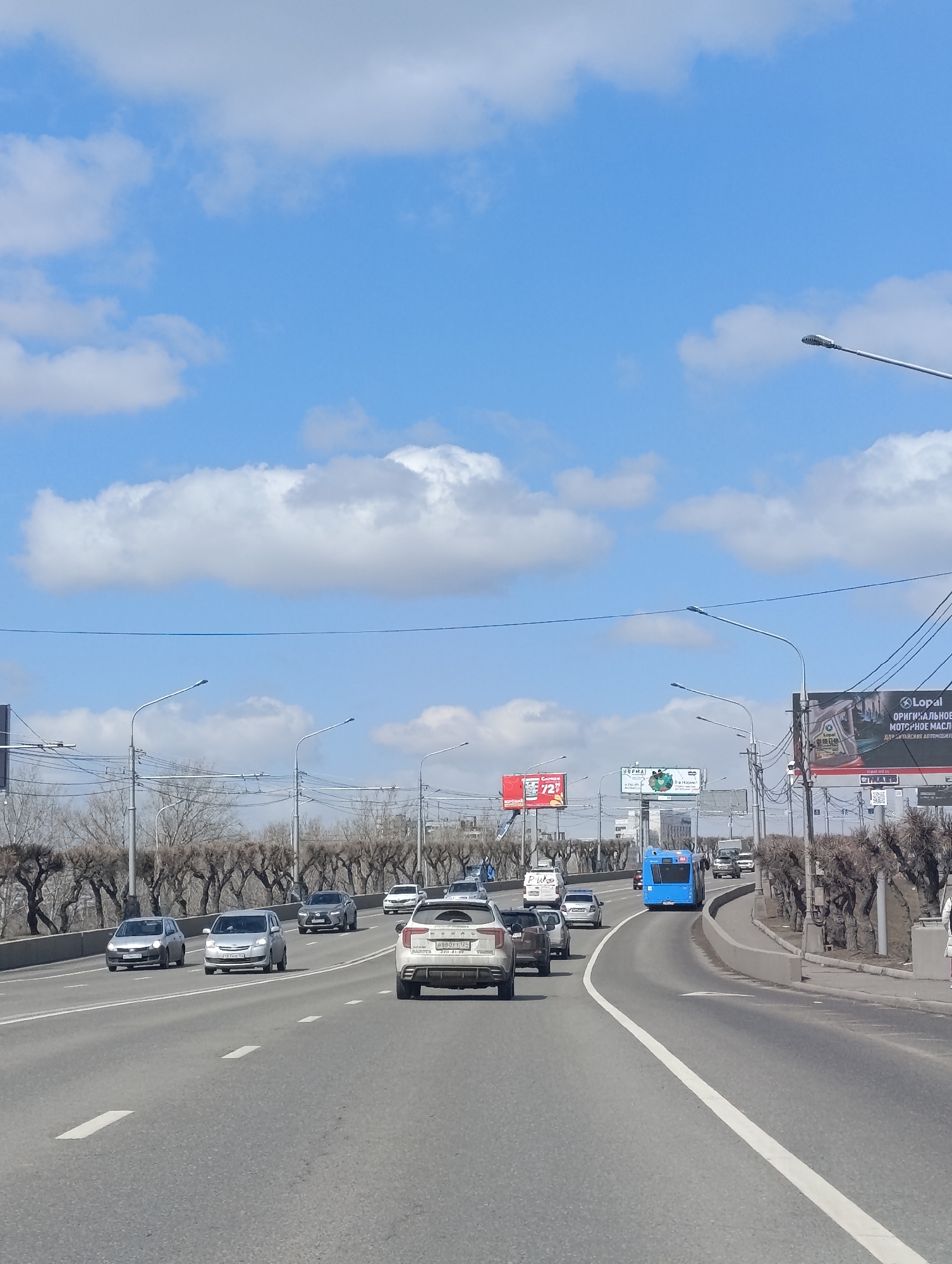 The image shows a multi-lane road in Krasnoyarsk, Russia, with moderate traffic consisting of cars and a blue bus. The road has a white line dividing the lanes. Bare trees line the side of the road, and billboards are visible in the distance. The sky is blue with scattered white clouds. Streetlights and power lines are also present.