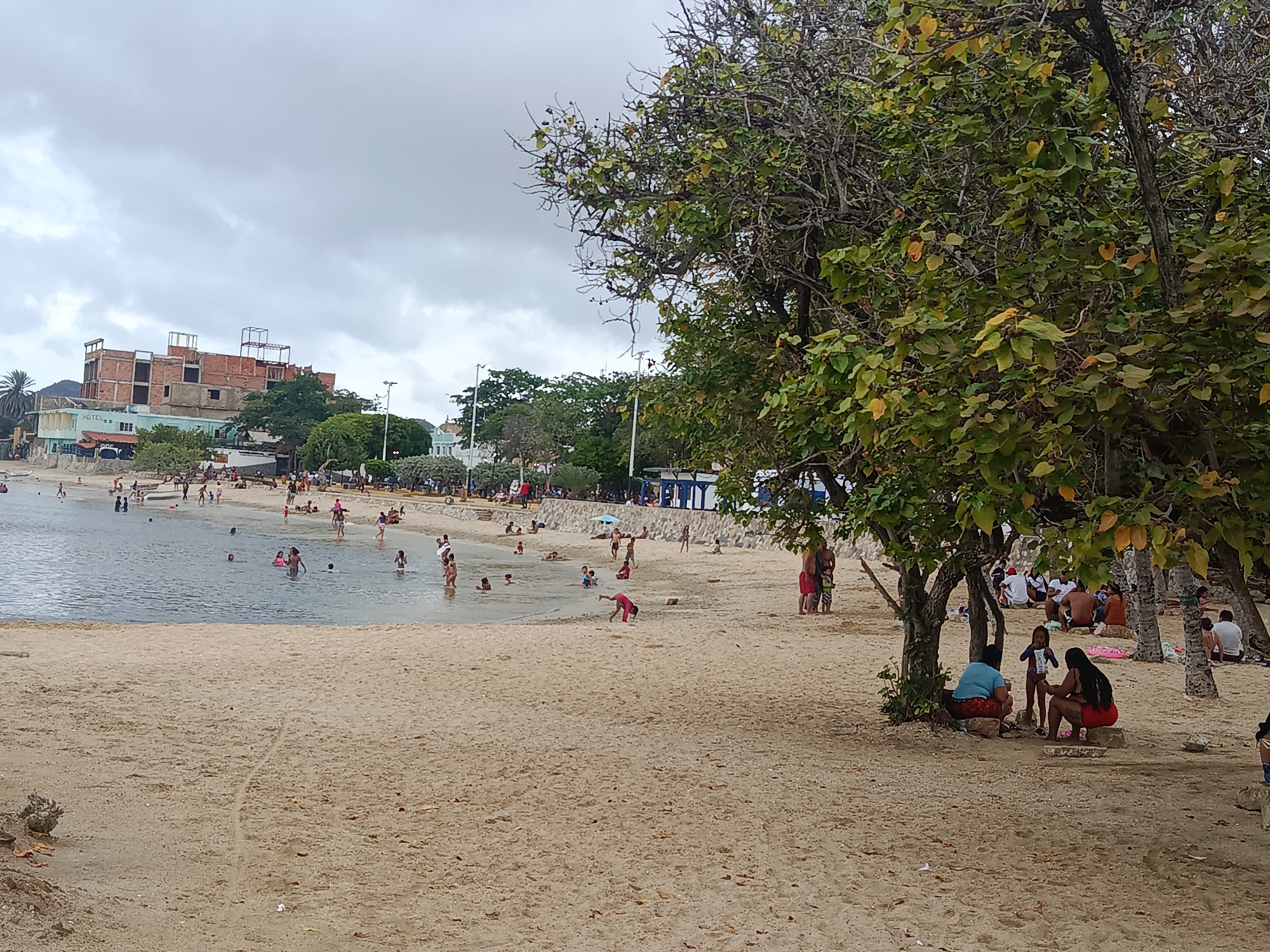 This image depicts a busy beach scene in Juan Griego, Venezuela, on an overcast day. The sky is dominated by a uniform layer of grey clouds.
In the foreground, a wide expanse of light brown sand stretches across the bottom half of the frame. Scattered small debris and faint tracks are visible on the sand. Towards the right, under the canopy of large, leafy trees, several individuals and small groups are seated or resting on the sand, some near the bases of palm trees. Two adults, one in a blue top and the other in red shorts, are prominently seated on the sand in the mid-right foreground, with a child standing between them.
The midground features a calm, shallow body of light blue-grey water where numerous people, including adults and children, are wading, swimming, or standing. Along the sandy edge of the water, more people are visible, with one child in a pink outfit captured in motion, bending forward on the sand. A grey stone or concrete retaining wall delineates the back of the beach, separating it from an elevated paved area.
In the background, behind the retaining wall and dense green foliage, several structures are visible. A multi-story building, appearing to be under construction, features exposed red brickwork and rebar extending upwards, indicating multiple unfinished floors. To its left, a completed light-colored building with the word "HOTEL" visible on its facade stands closer to the water. Lush green trees and other vegetation are abundant, lining the coastline and interspersed among the buildings. Several streetlights are also visible along the elevated area. Distant hills or mountains are faintly discernible on the horizon to the far left.