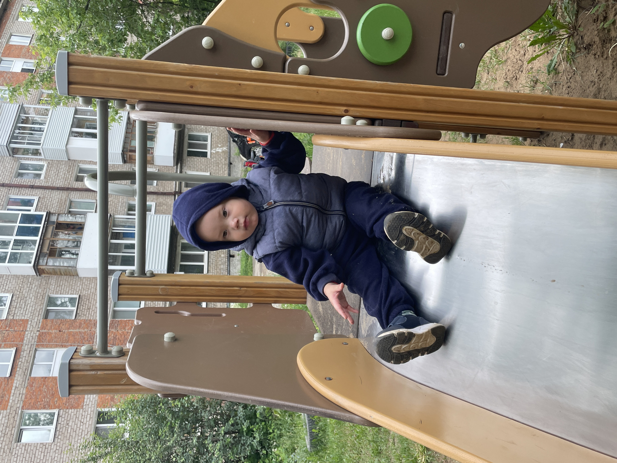 A young child, appearing to be a toddler of East Asian descent, is seated on a silver-colored metal slide within a playground structure. The child is dressed in a dark blue hooded tracksuit, a grey sleeveless puffer vest, and dark athletic shoes with white soles. The child's right hand is extended towards the light brown plastic side support of the slide, while the left hand rests on the metallic sliding surface. The child's gaze is directed slightly to the left.
The playground equipment features light brown plastic components, including the slide's frame, and vertical wooden-look posts. A green circular disc with a white center cap is visible on the right side of the structure, and grey metal bars are positioned above the child.
In the background, a multi-story brick apartment building with a mix of light-colored and reddish brickwork is prominent. Numerous white-framed windows and several enclosed or partially enclosed balconies with white or grey siding are visible on the building. Green trees and bushes are present on either side of the playground and further in the background. The ground around the playground is a mix of bare soil and sparse green grass. This scene is located in the city of Perm, Russia.