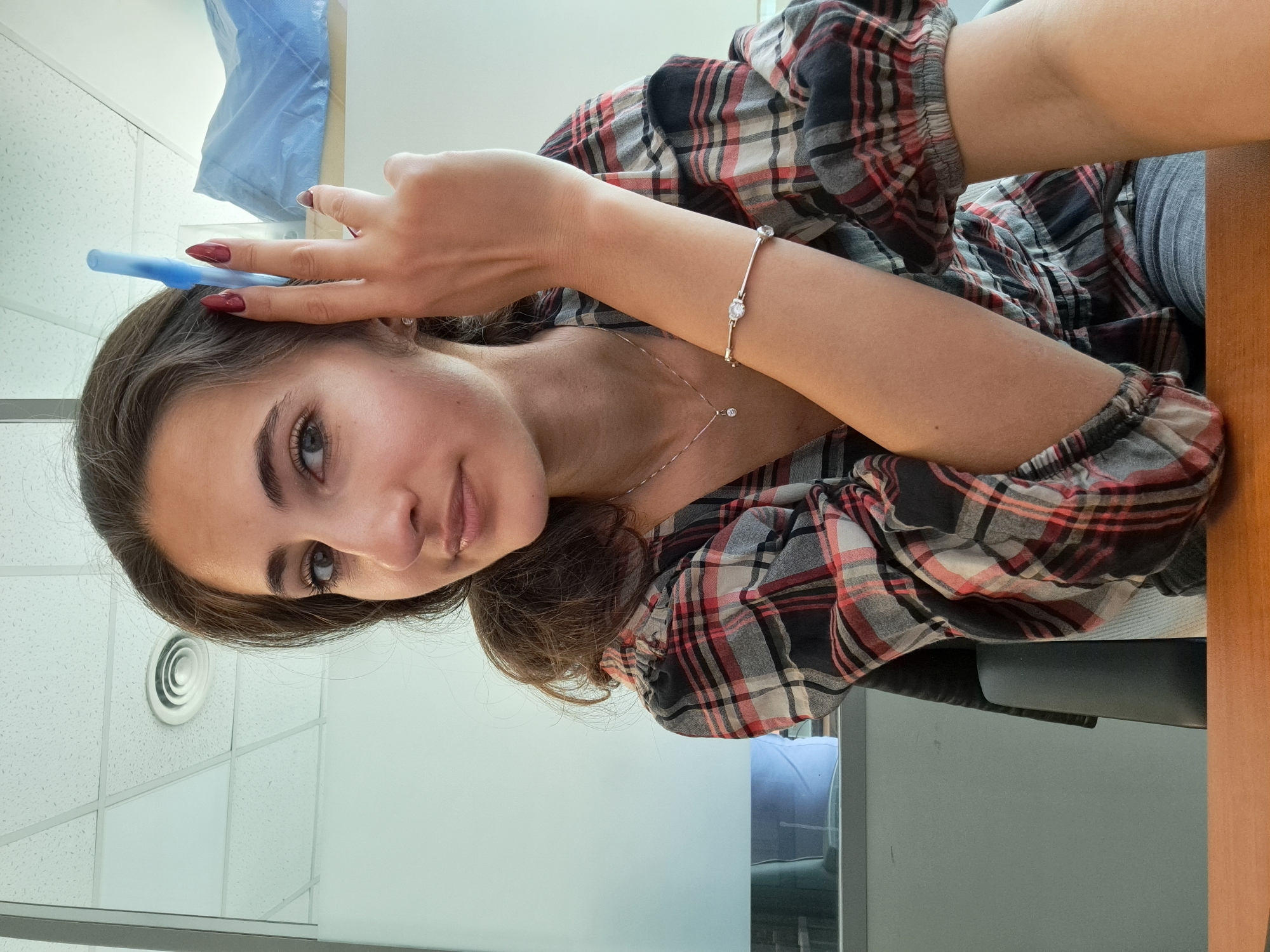 A young woman with brown hair and blue eyes is indoors in what appears to be an office setting. She is wearing a plaid shirt and has a bracelet on her left wrist. She holds a blue, possibly plastic, object near her forehead with her left hand, and her nails are painted dark red. The background includes a white ceiling with circular vents, a window, and what appears to be a blue medical drape or covering. There's a desk or table visible in the lower right corner of the frame. There is no immediate evidence to suggest the image was taken in Sofia, Bulgaria. The description is limited to the available visual information.