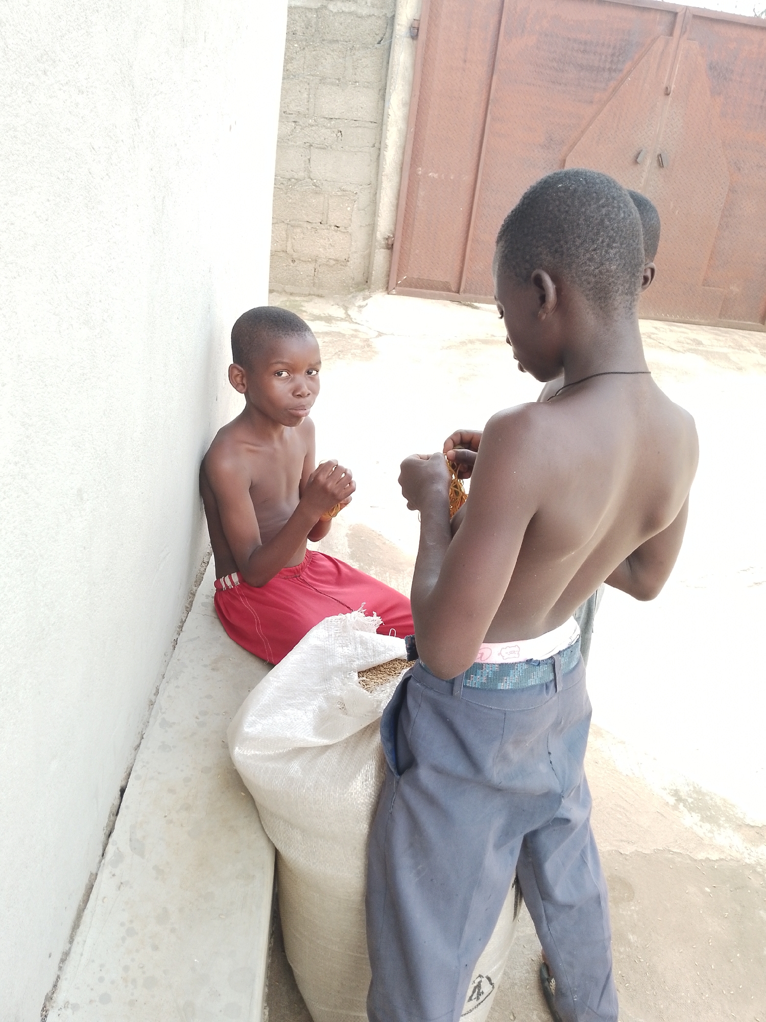 Three young boys are shown outdoors in what appears to be a casual, urban environment in Balewa, Nigeria. A boy in red shorts is seated against a white wall, holding something in his hands. To his right, another boy in gray pants stands and examines something in his hands, which appears to be food items. A third boy's head is visible behind the standing boy, partially obscured. A large, off-white sack is positioned in front of the seated boy, containing grain. The background features a rough, textured wall and a large, reddish-brown metal gate. The ground is paved with concrete.