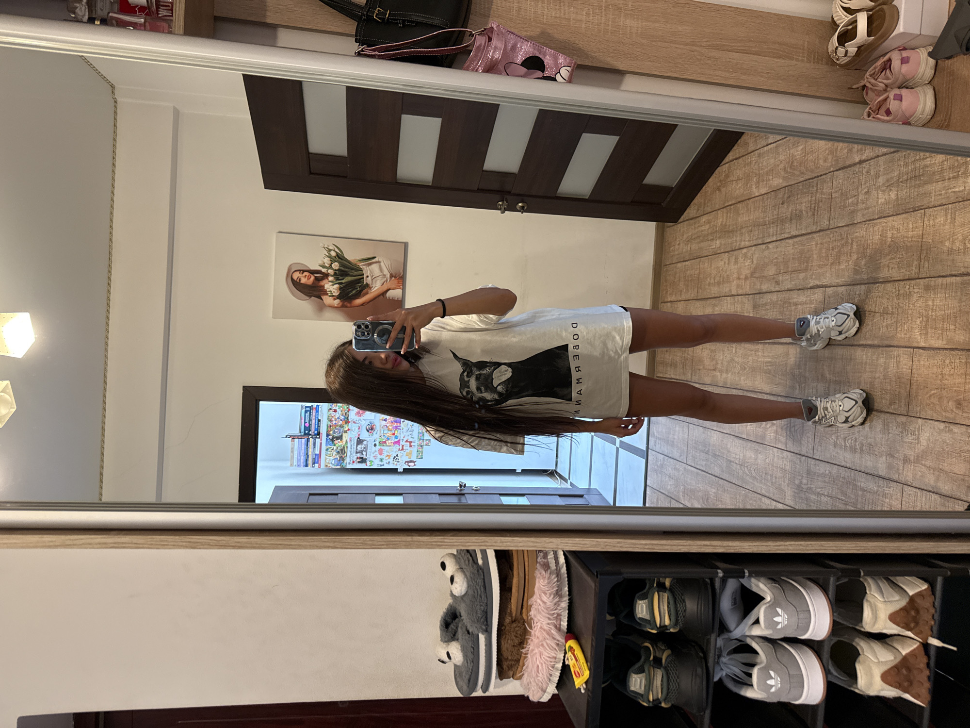 A young woman with long dark hair is taking a mirror selfie in what appears to be a bedroom or dressing area. She is wearing a white t-shirt with a black dog graphic and text, and dark shorts. She is holding a smartphone in her right hand. To her left, a multi-tiered shoe rack holds several pairs of shoes and slippers. To her right, a dark wooden door with rectangular inlays is visible, and on the wall next to it, a black handbag and a pink Minnie Mouse bag are hanging. A framed picture of a woman holding tulips hangs on the wall in the reflection of the mirror. The floor is tiled with a wood-grain pattern. The room has light-colored walls and a ceiling with a light fixture. The overall impression is of a personal space, likely in a residential setting in Chișinău, Moldova.