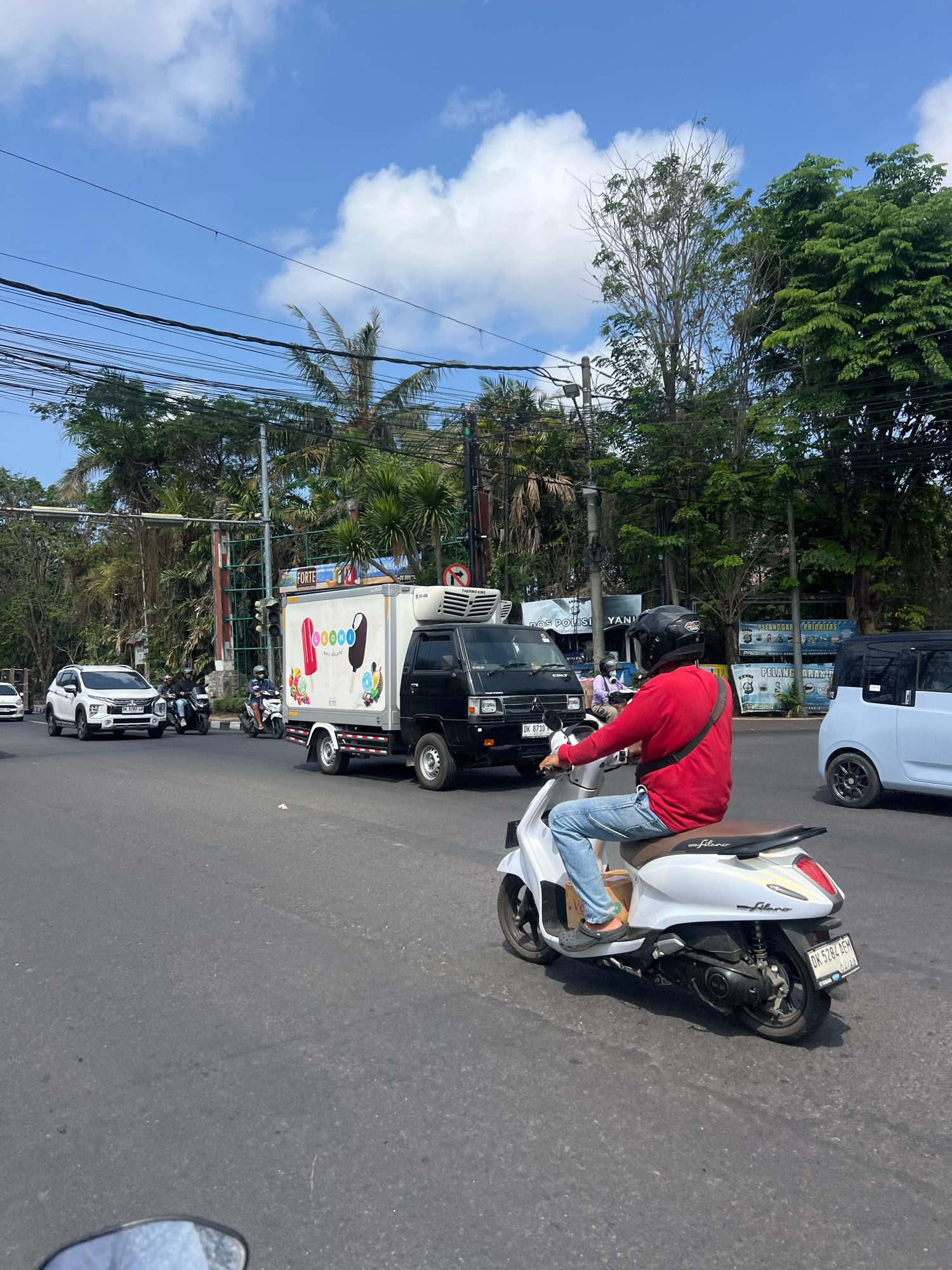 The image captures a street scene in Denpasar, Indonesia, on a bright, sunny day with scattered clouds. In the foreground, a person wearing a red long-sleeved shirt, blue jeans, and a black helmet is riding a white scooter. The scooter has a brown seat and a license plate that reads "DK 5284 AEM". The scooter is moving on an asphalt road, with its right side slightly angled towards the viewer.
Several other vehicles are present in the mid-ground. A black refrigerated truck with "FORTE" and colorful ice cream graphics on its side is driving in the same direction as the scooter. Next to the truck, a white SUV with license plate "DK 1287 LA" is also on the road. Further back, two people on a motorcycle are visible, and a light blue compact car is on the right side of the frame.
The environment is characterized by lush green trees lining the road and a clear blue sky overhead. Electrical wires are strung across the top of the image. Signs are visible in the background, including one that says "PELANGGARAN PRIORITAS" (Violation of Priority). The overall impression is of a busy urban street in a tropical city.