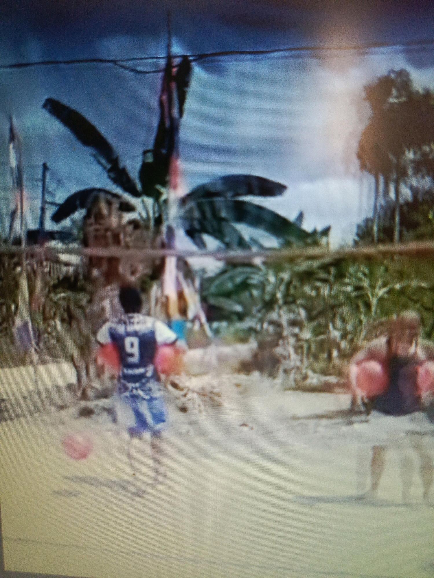 The image depicts an outdoor scene with two individuals engaged in an activity, possibly a game, on a dirt or sand-covered surface. In the foreground, a person wearing a blue and white jersey with the number "9" on the back is holding two red objects, one in each hand, and looking towards the right. This individual is also wearing blue shorts. A pink sphere, likely a ball, is visible on the ground to the left of this person. In the background, another person, partially obscured and appearing blurry, is also holding red objects. The environment suggests a warm climate with lush green foliage, including what appear to be banana trees, and some structures in the distance. The overall impression is of a casual gathering or a sporting event in a rural or semi-rural setting, possibly in Tik Jeniak, Indonesia, given the vegetation and lighting. The sky is overcast with a blueish hue.