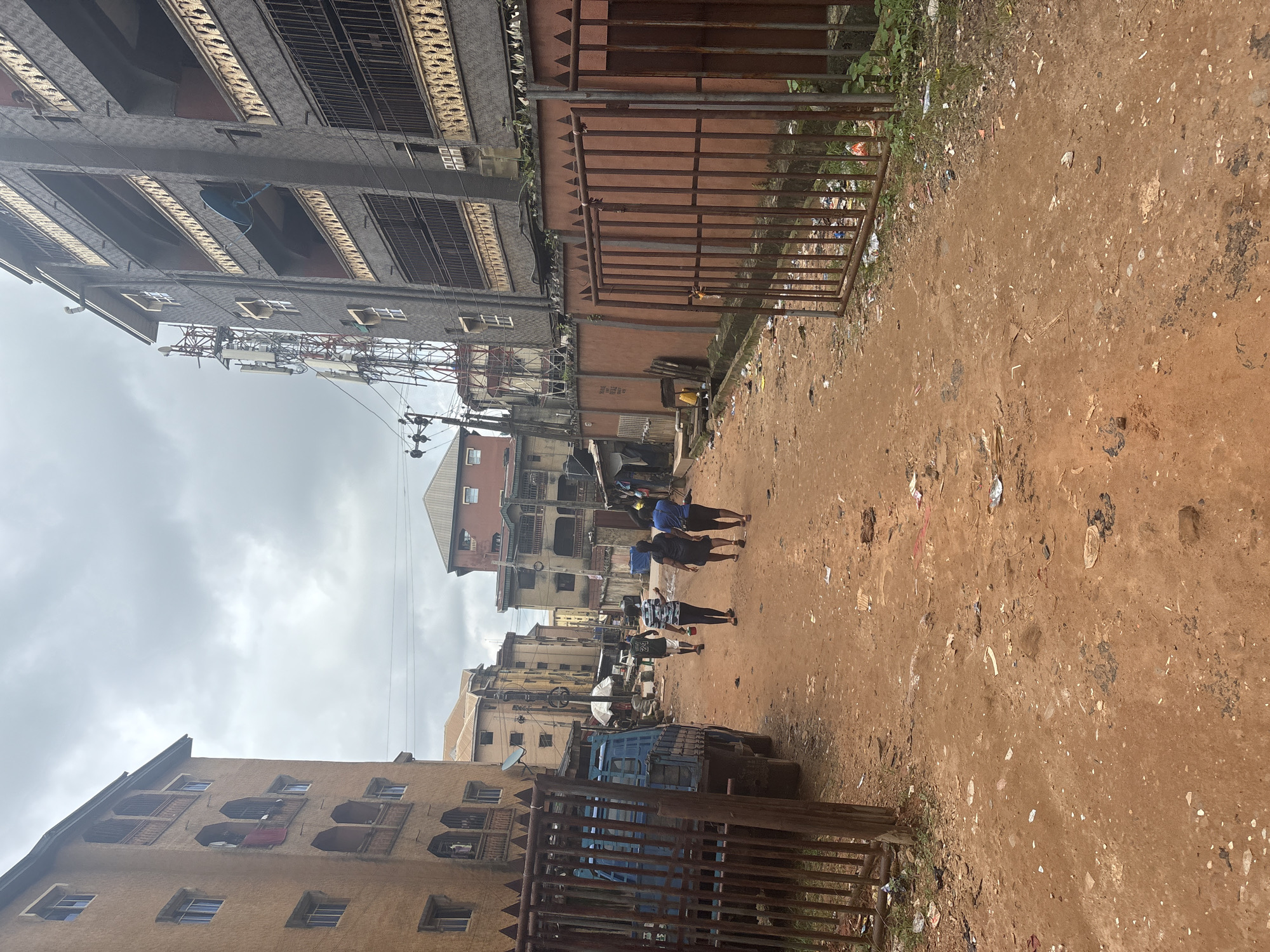 In Onitsha, Nigeria, a dusty, reddish-brown dirt road winds between dense residential buildings. The buildings, some appearing aged with balconies and numerous windows, line both sides of the narrow street, creating an enclosed urban atmosphere. A blue truck is parked on the left, partially obscured by a rusted metal gate. On the right, a similar rusted gate guards a property with a low, reddish-brown wall. Several individuals, including three women and a man, walk away from the viewer down the road. The sky above is overcast with thick, grey clouds. Scattered litter is visible on the road and along the sides, indicative of an urban environment. The scene suggests a typical day in a densely populated area of Onitsha.