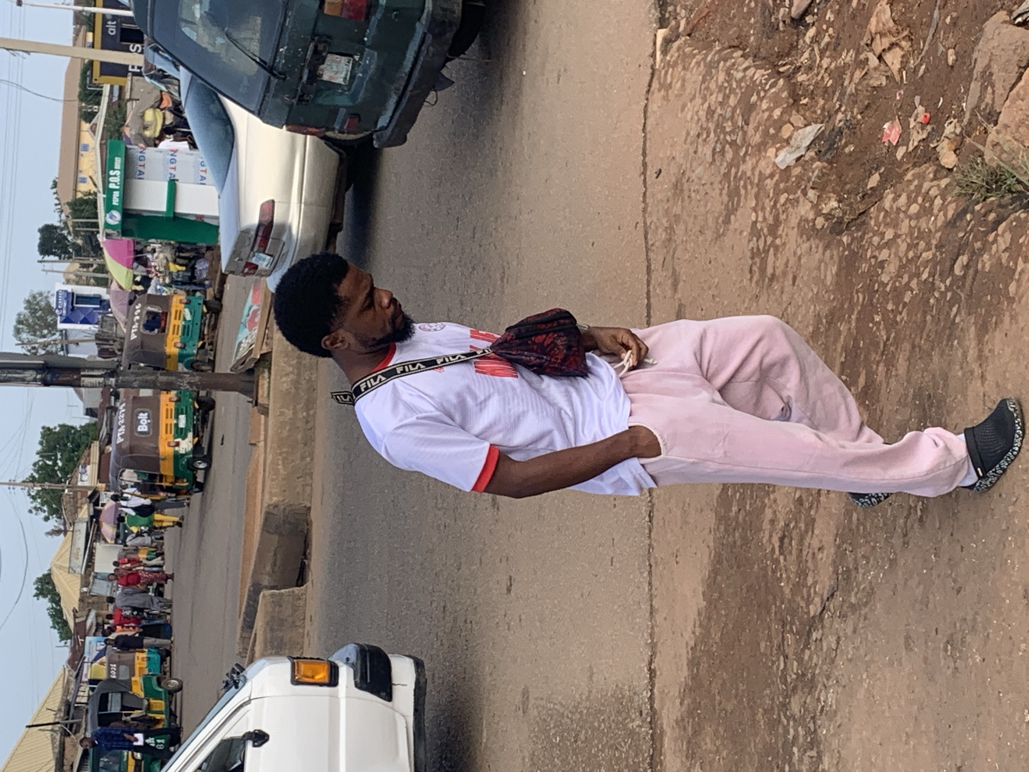 The image depicts a street scene in Gura Top, Nigeria. In the foreground, a young Black male is walking and looking down. He is wearing a white t-shirt with red trim, pink sweatpants, and black clogs. A small, patterned bag is slung across his chest. In the background, several auto-rickshaws, commonly known as "Keke Napep," are parked or driving. These vehicles are yellow and green, some with "Bolt" branding. A silver sedan and a dark green van are also visible in the street. Shops with various wares and umbrellas are set up along the roadside. The ground is a mix of asphalt and dirt, with some debris present. The overall environment suggests a bustling, everyday market or transport hub.