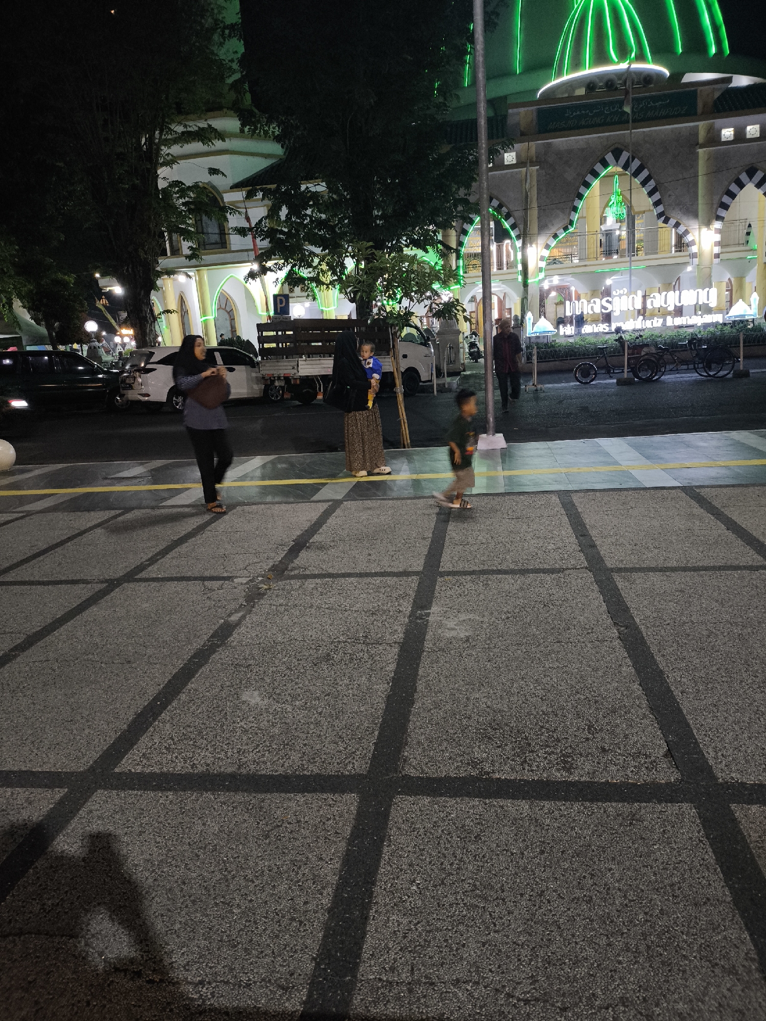 The image depicts an evening scene outside the Grand Mosque KH. Anas Machfuz in Lumajang, Indonesia. The mosque is illuminated with vibrant green neon lights, highlighting its domes and arched windows. In the foreground, a pedestrian walkway with a distinctive grid pattern is visible.
Several people are present. A woman wearing a hijab and a long skirt walks across the pedestrian area, carrying a dark bag. Beside her, another person, possibly a woman in a dark hijab, holds a child who is wearing a blue shirt. To the right, a young boy in a dark shirt and light-colored shorts walks on the pedestrian walkway, facing away from the camera. A man in a maroon shirt is walking in the background near a parked truck.
Several vehicles are parked along the street, including a white truck and a white car. Two bicycles are parked near the mosque's entrance. The environment appears to be a well-lit urban area at night, with trees lining the street and contributing to the overall atmosphere. The text on the mosque reads "MASJID AGUNG KH. ANAS MACHFUZ LUMAJANG".