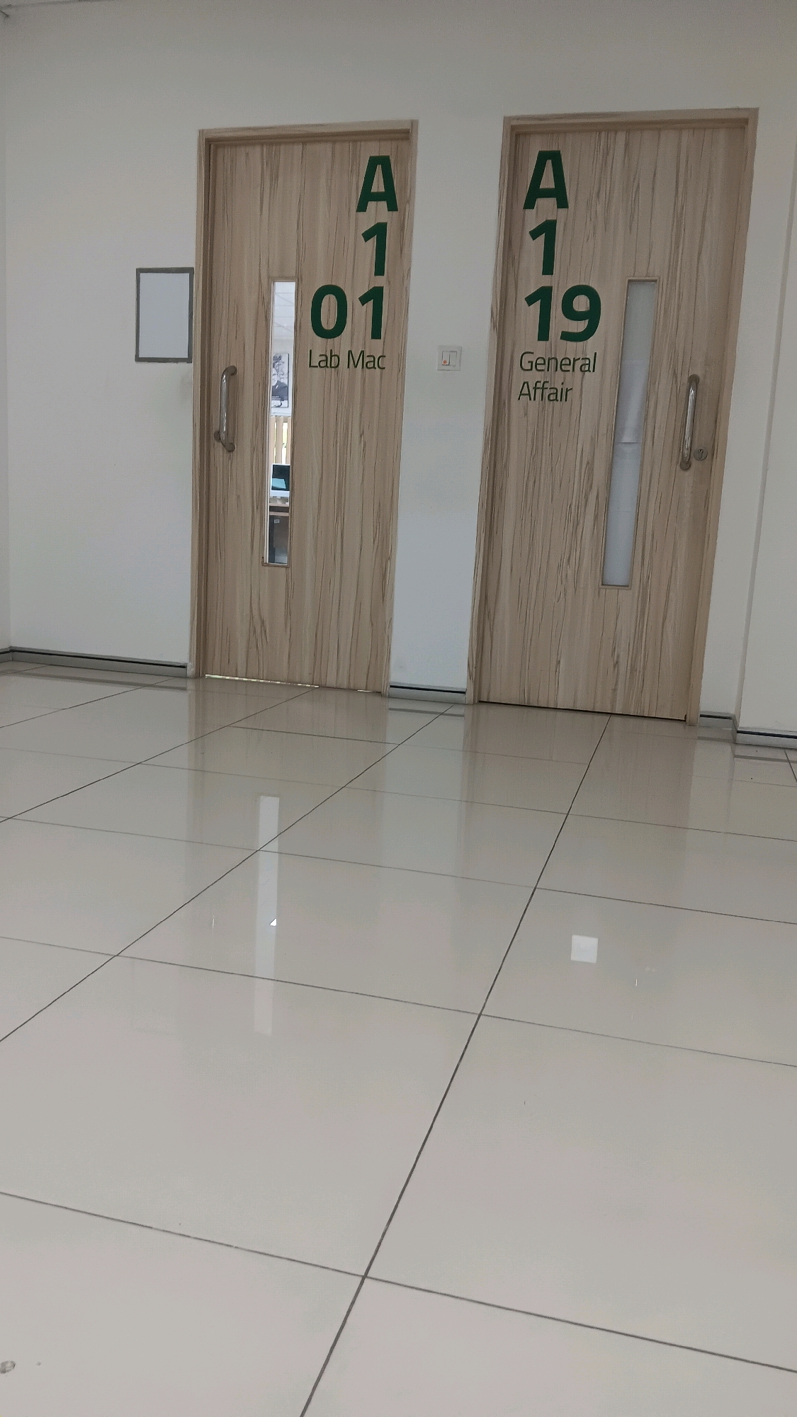 Two wooden doors with vertical grain patterns are positioned side-by-side in a well-lit hallway with a tiled floor. The door on the left is labeled with green text: "A 1 01" and below that, "Lab Mac". This door has a glass pane with a reflective interior and a silver handle. The door on the right is labeled with green text: "A 1 19" and below that, "General Affair". This door also has a glass pane and a silver handle. To the left of the left door, a small white rectangular sign is mounted on the wall. The floor is covered in large, light-colored square tiles with dark grout lines, reflecting the overhead lighting. The environment appears to be an indoor public space, possibly a hospital, clinic, or administrative building. The image does not provide any information about the city of Tangerang, Indonesia.