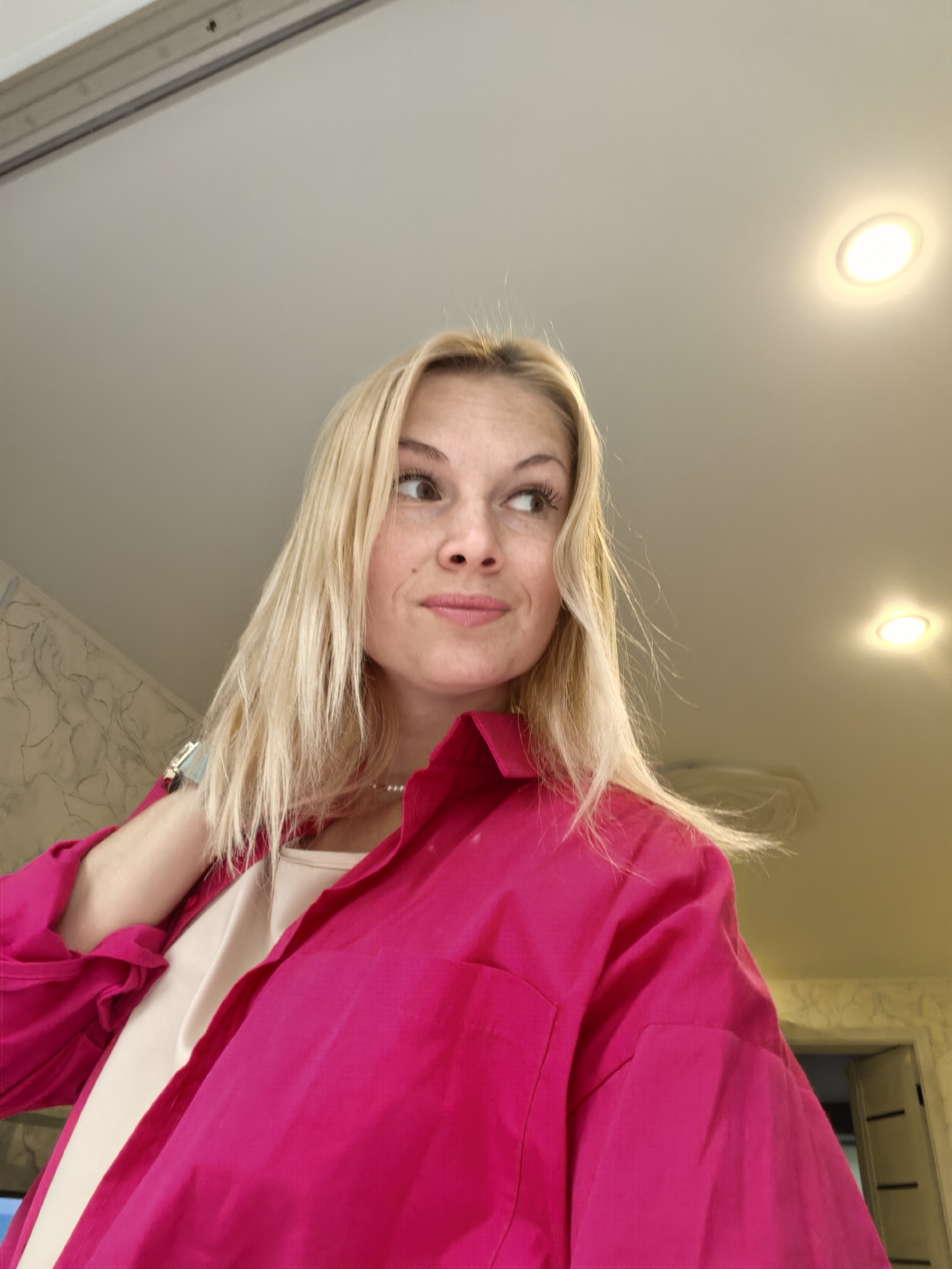 A blonde woman is looking off to the side. She is wearing a bright pink button-up shirt over a beige tank top and a pearl necklace. The background appears to be indoors, with a ceiling light visible. The overall setting does not provide enough information to determine a specific location within Sterlitamak, Russia.