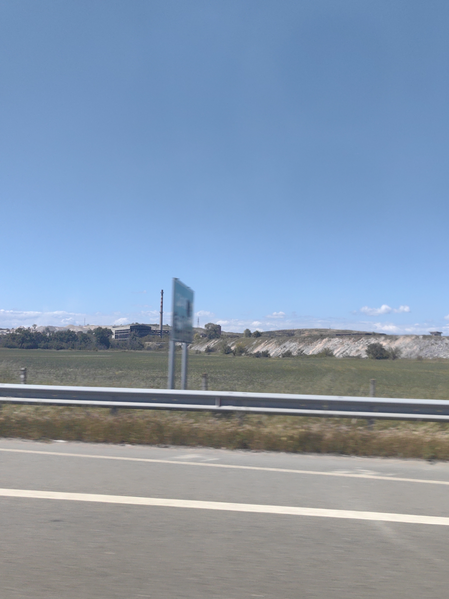 A daytime roadside view in or near Belozem, Bulgaria. In the distance are buildings and a smokestack, likely industrial facilities, positioned on the horizon against a clear blue sky. A faded or blurred sign stands near the guardrail, obscuring details. A grassy field and some elevated terrain are visible between the road and the buildings. The road stretches across the foreground.