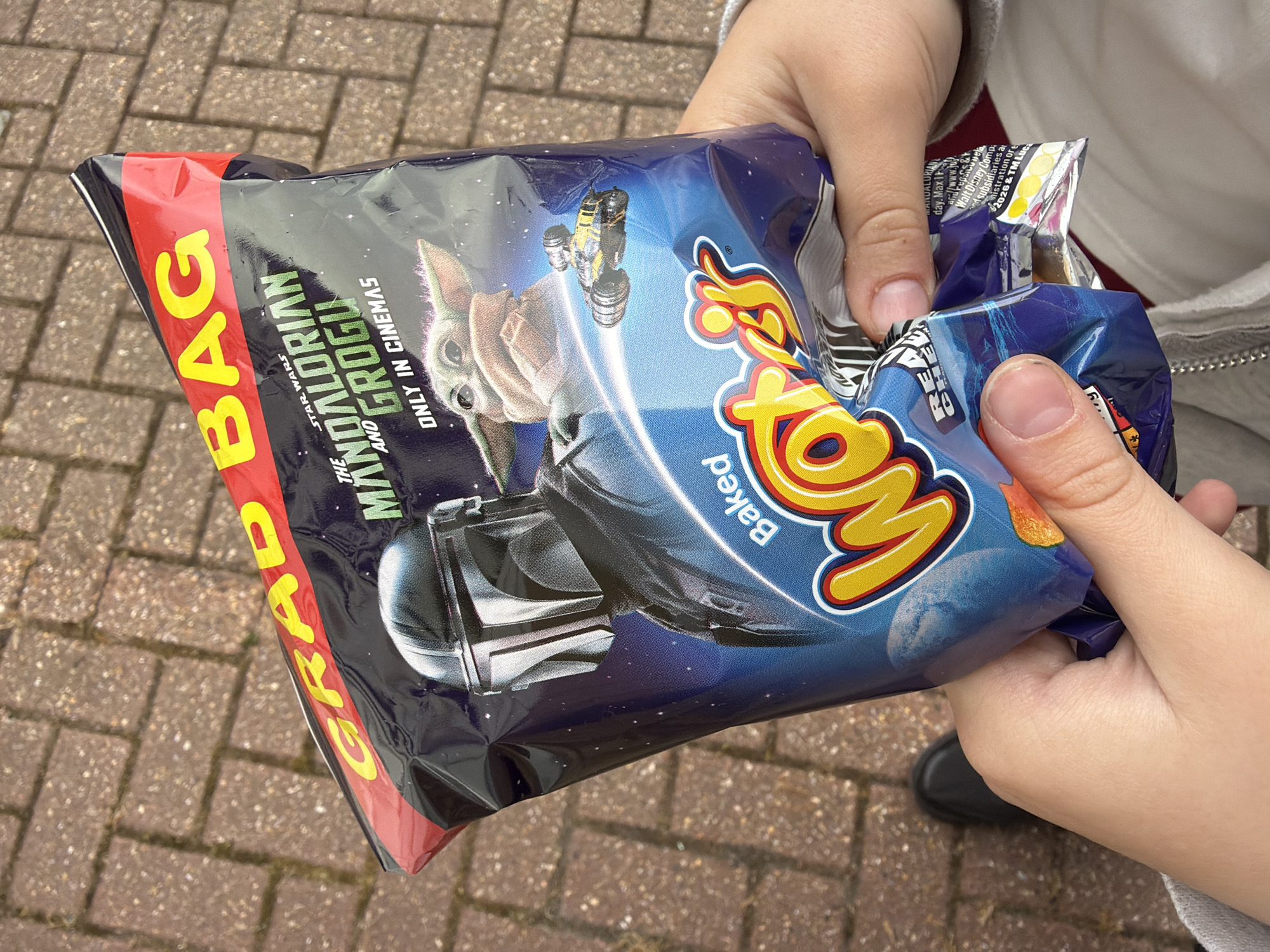 A person's hands hold a half-open bag of "Wotsits Baked" crisps, featuring "Star Wars: The Mandalorian and Grogu" branding. The bag, labeled "GRAD BAG" in red and yellow, displays an image of The Mandalorian's helmet and Grogu. The background is a brick-paved ground. The person is wearing a light grey sweater. A glimpse of a black shoe is visible near the bottom right of the frame.