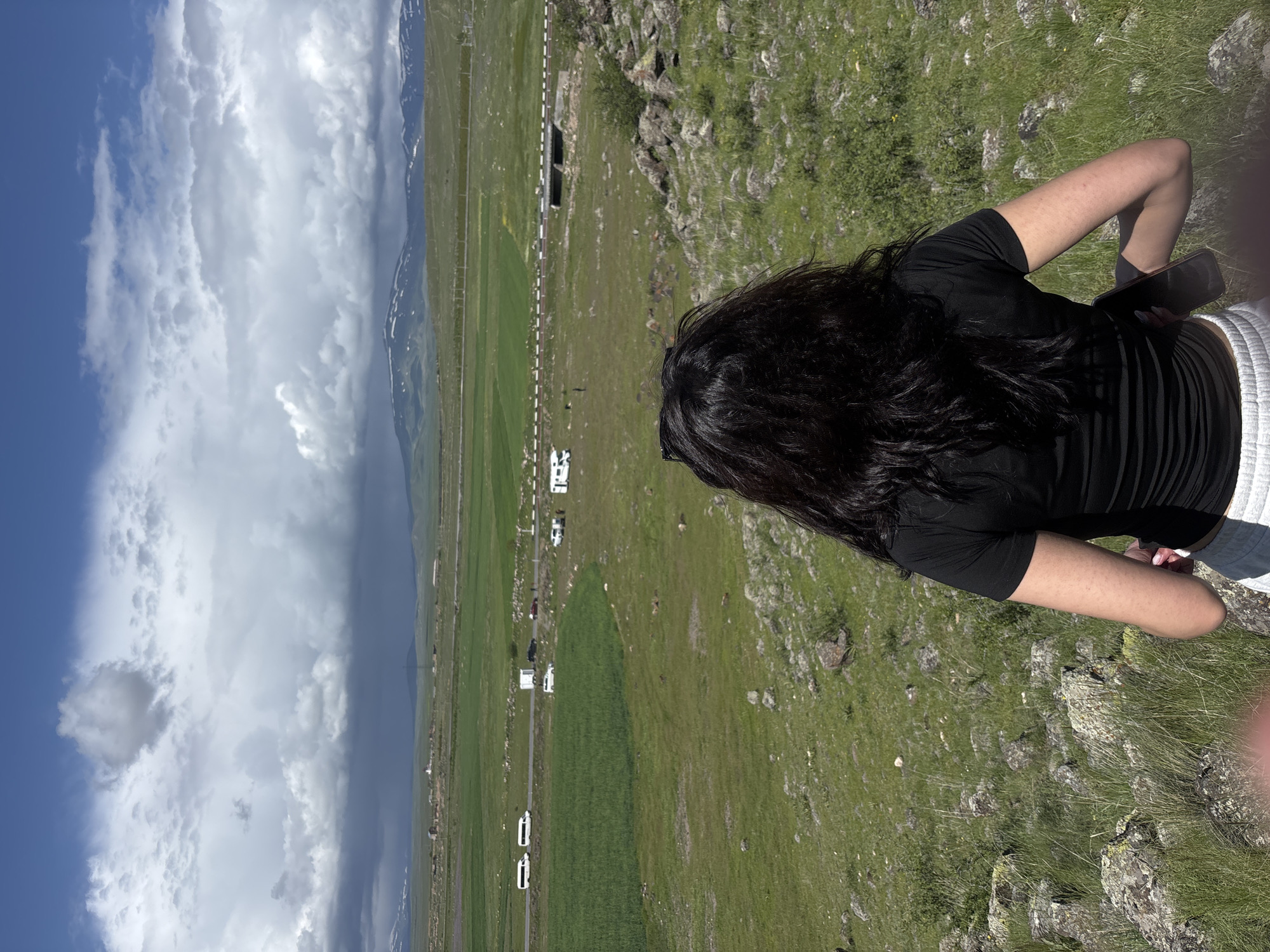 A medium, angled outdoor shot shows a person with long, dark hair wearing a black t-shirt, gray shorts, and holding a black phone, looking out over a green, grassy landscape. In the background, there is a road with several white RVs parked along it, a field of green crops, and mountains in the distance. The sky is blue with white clouds. The person is standing on a rocky area with sparse vegetation. Based on the landscape and the presence of RVs, this scene could plausibly be located near Tlik, Armenia.