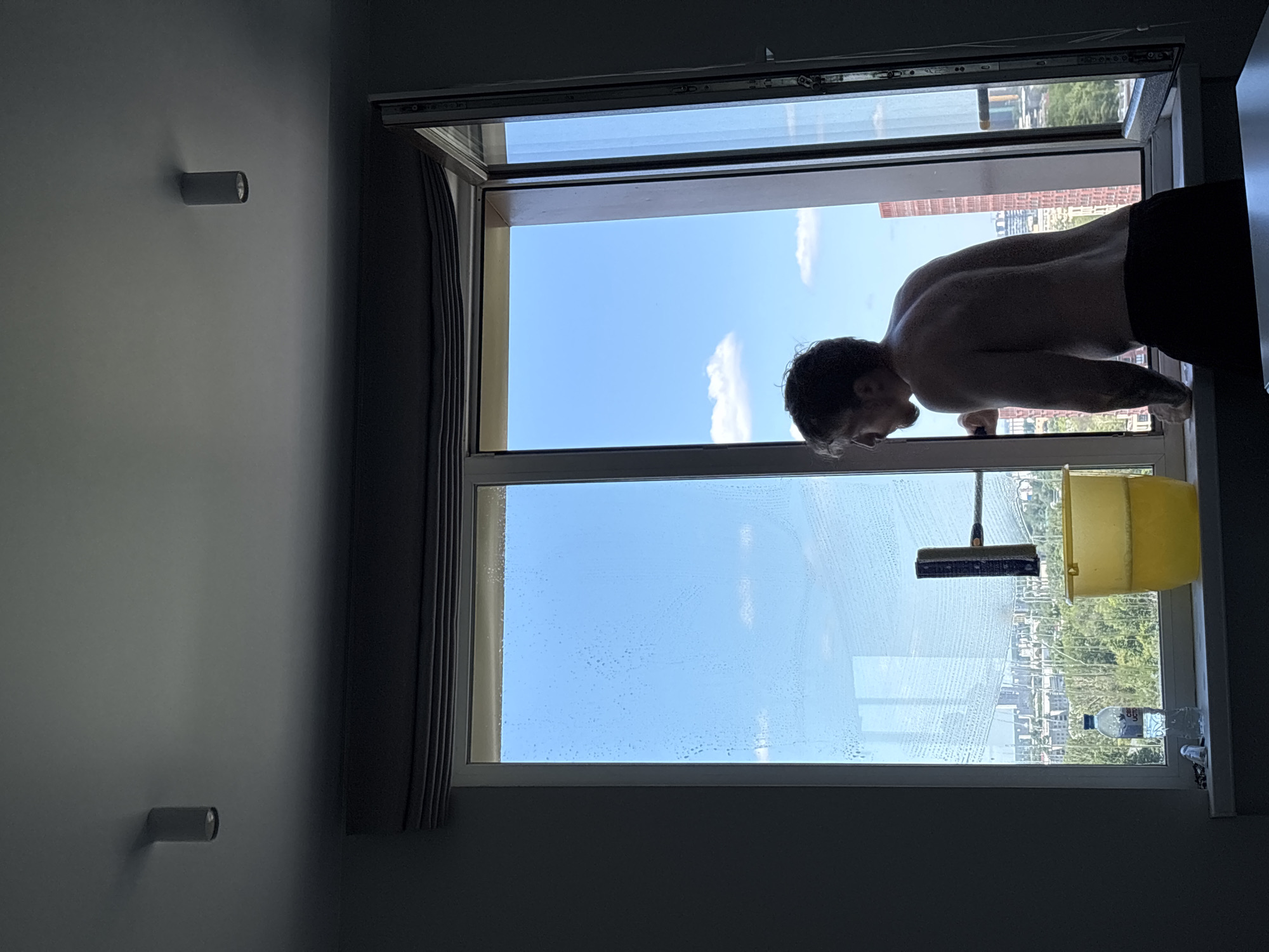 The image displays an interior scene looking out through a window where a man is cleaning. The ceiling is light grey, equipped with two dark, cylindrical flush-mounted lights—one on the left and one on the right. Dark grey window blinds are retracted at the top of the window frame. The interior walls are dark grey.
A shirtless man, with light brown hair and a beard, is silhouetted as he stands facing right and slightly away from the viewer. He wears black shorts or trousers and exhibits tattoos on his right forearm. He holds a window squeegee with a dark cleaning head and an orange handle, actively applying it to the left window pane, which shows water droplets and streaks of soap.
On the white window sill are a clear plastic water bottle with a blue label on the left, a yellow plastic bucket containing yellowish soapy water in the center, and a small white object, possibly a cloth, near the water bottle.
The window structure includes multiple panes. The left pane is wet with cleaning solution. The rightmost pane is open inward, revealing its frame, hinges, and locking mechanisms.
Beyond the window, a clear blue sky with scattered white cumulus clouds is visible. The outdoor environment, identified as a cityscape in Nizhny Novgorod, Russia, shows a substantial amount of green foliage and trees in the mid-ground. In the background, urban buildings are present, notably a tall, reddish-brown, multi-story building with a rectilinear design visible through the open window pane on the right, with another similar structure partially obscured behind it.