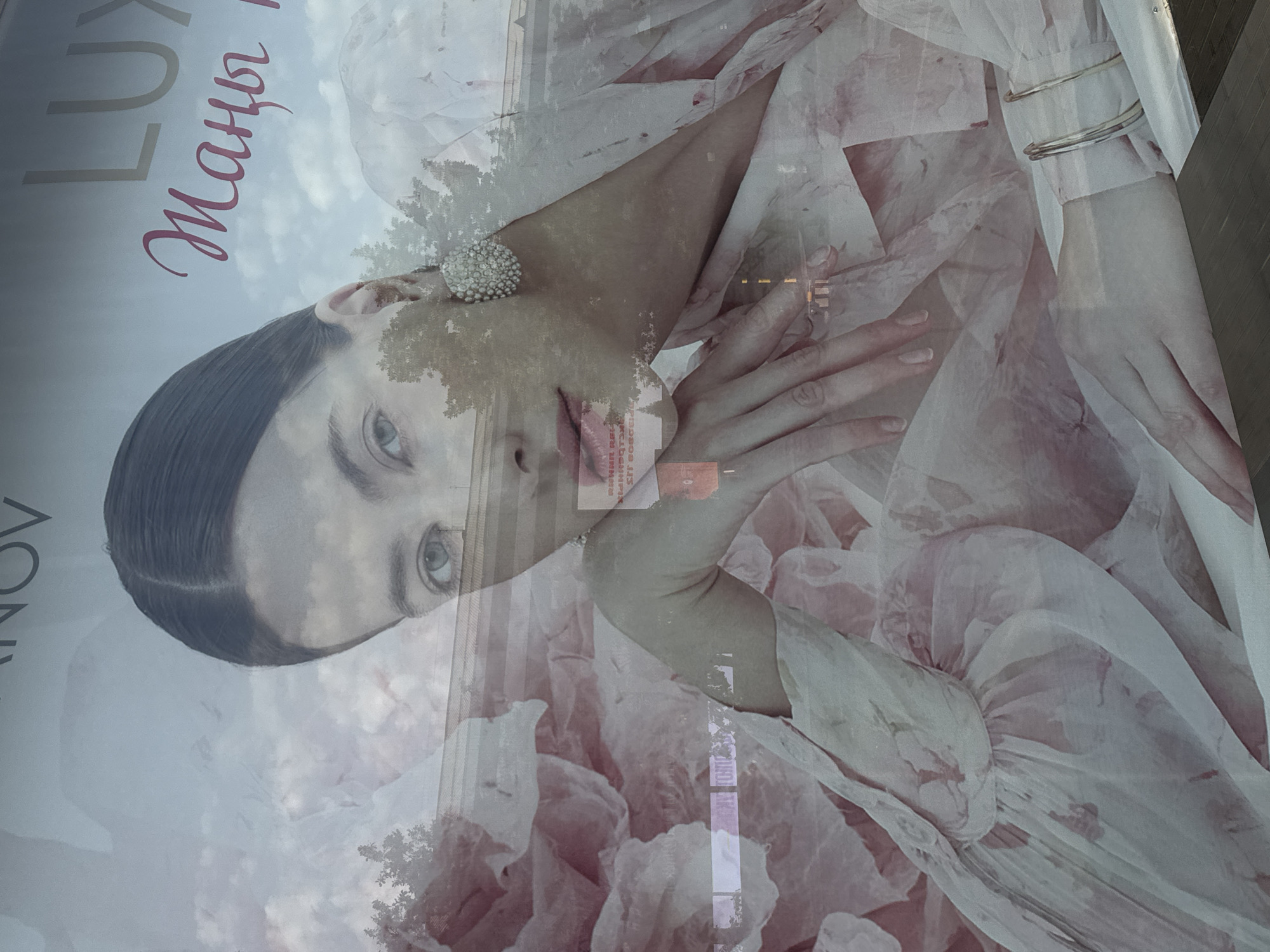 The image is a close-up, highly reflective outdoor photograph of a billboard advertisement featuring a woman. The woman is shown from the chest up, with her head turned to her right. She has dark hair pulled back and is wearing a large, ornate earring on her right ear. Her lips are pink. The billboard itself has text, with "LUX" visible in the upper right corner and what appears to be Cyrillic or a similar script in pink below it. The advertisement is layered with reflections of the sky, clouds, trees, and buildings, suggesting it is mounted on a glass surface in an urban environment. The overall color palette is soft, with pastel pinks, whites, and muted blues from the sky. This billboard is located in Bishkek City, Kyrgyzstan, and is likely situated along a busy street or thoroughfare, as indicated by the reflections of traffic and city infrastructure.