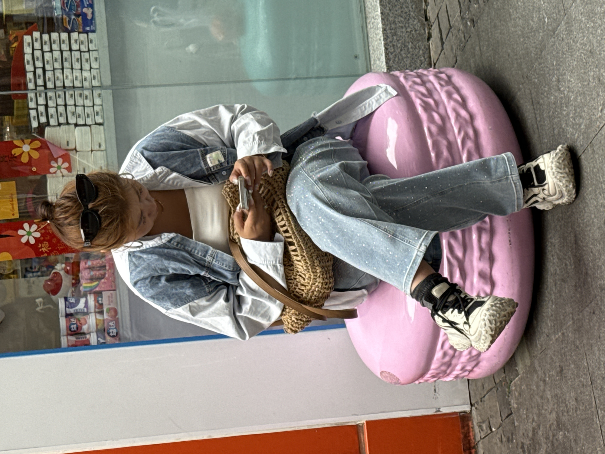 A young woman sits on a pink, rounded outdoor bench in the Yuzhong District, China, near a storefront. The storefront's window displays products, including boxes of tissues and candies. The woman wears a denim jacket over a white top, sparkly blue jeans, and white sneakers with black laces. She holds a small device, possibly a phone, in her hands. A straw bag rests beside her. The ground next to the bench is paved with gray stone.