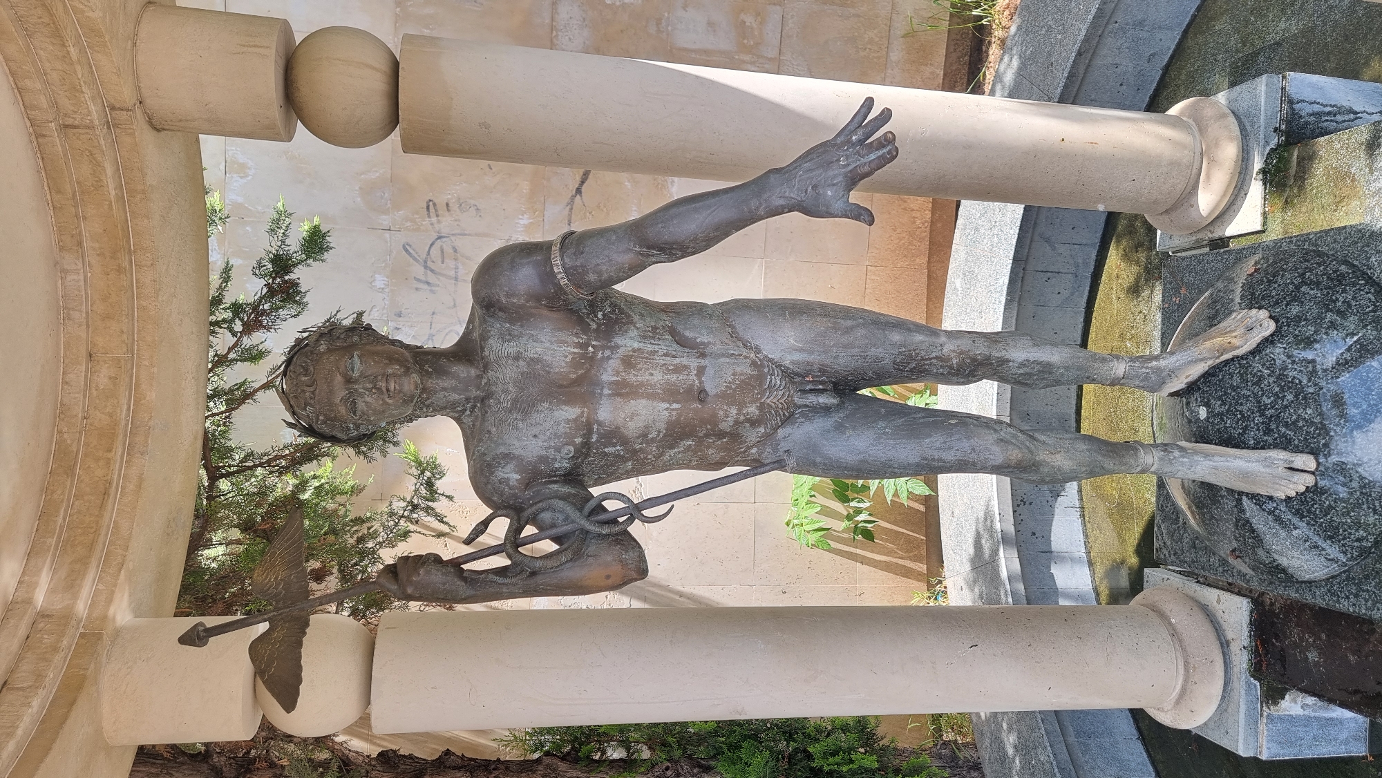 The image depicts a bronze statue of a nude male figure located in Sofia, Bulgaria. The statue appears to be part of a fountain or water feature. The figure is posed with one arm raised and the other holding a staff entwined with a snake, potentially representing Asclepius. The statue is surrounded by architectural elements including columns and stone walls. Some vegetation, including trees and smaller plants, is also visible. The statue stands atop a dark stone pedestal with water flowing around its base. The overall setting suggests a public space, likely a park or square within the city.
