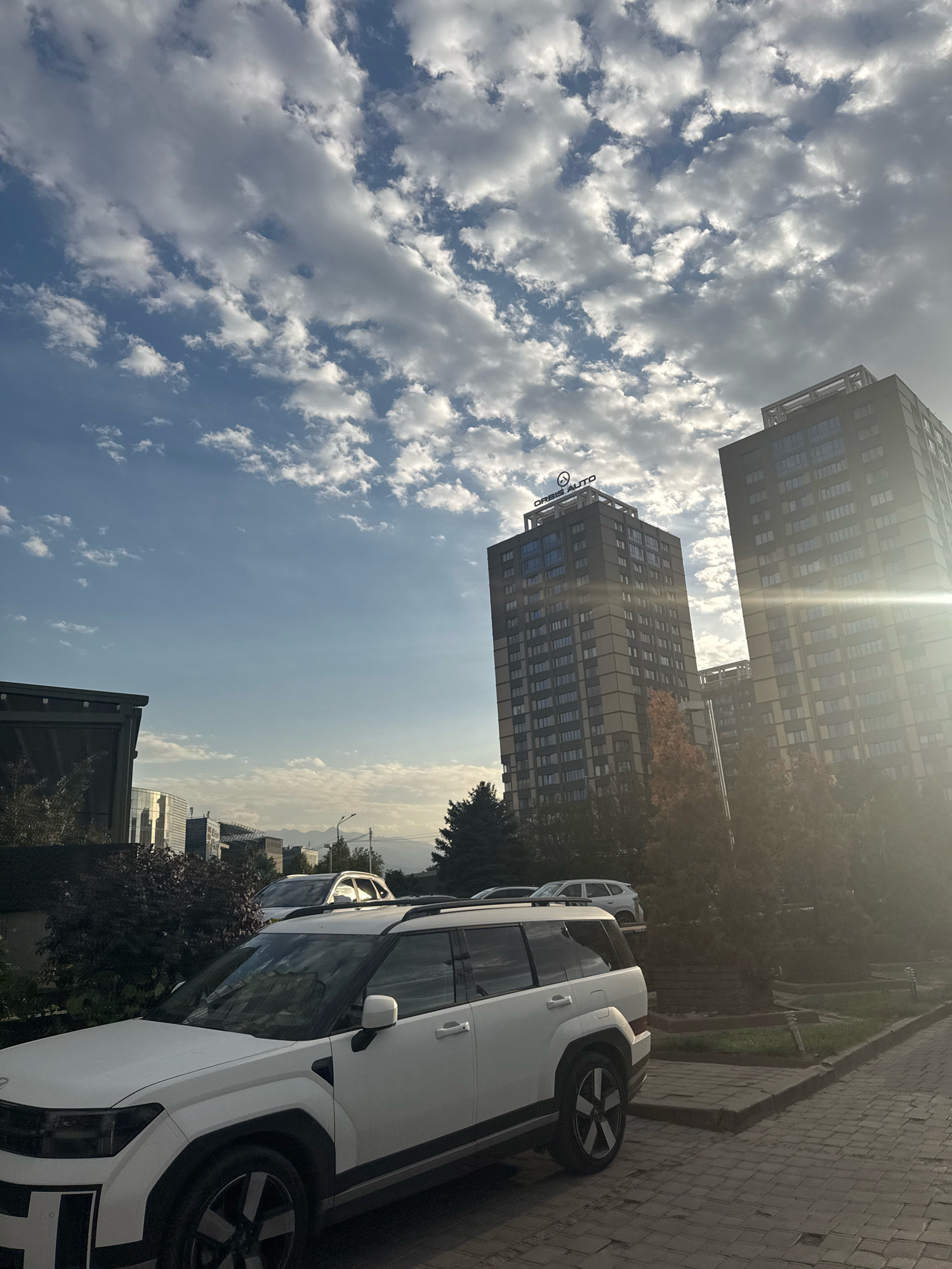 The image depicts a street scene in the city of Almaty, Kazakhstan, during daytime. The foreground features a white SUV parked on a cobblestone street. In the mid-ground, several trees and a dark building are visible. The background is dominated by two tall, modern apartment buildings with numerous windows. The building on the left has a sign at its top that reads "ORBIS AUTO". The sky is blue with scattered white clouds, and the sun is shining brightly, causing lens flare on the right side of the image. Mountains can be seen in the far distance.