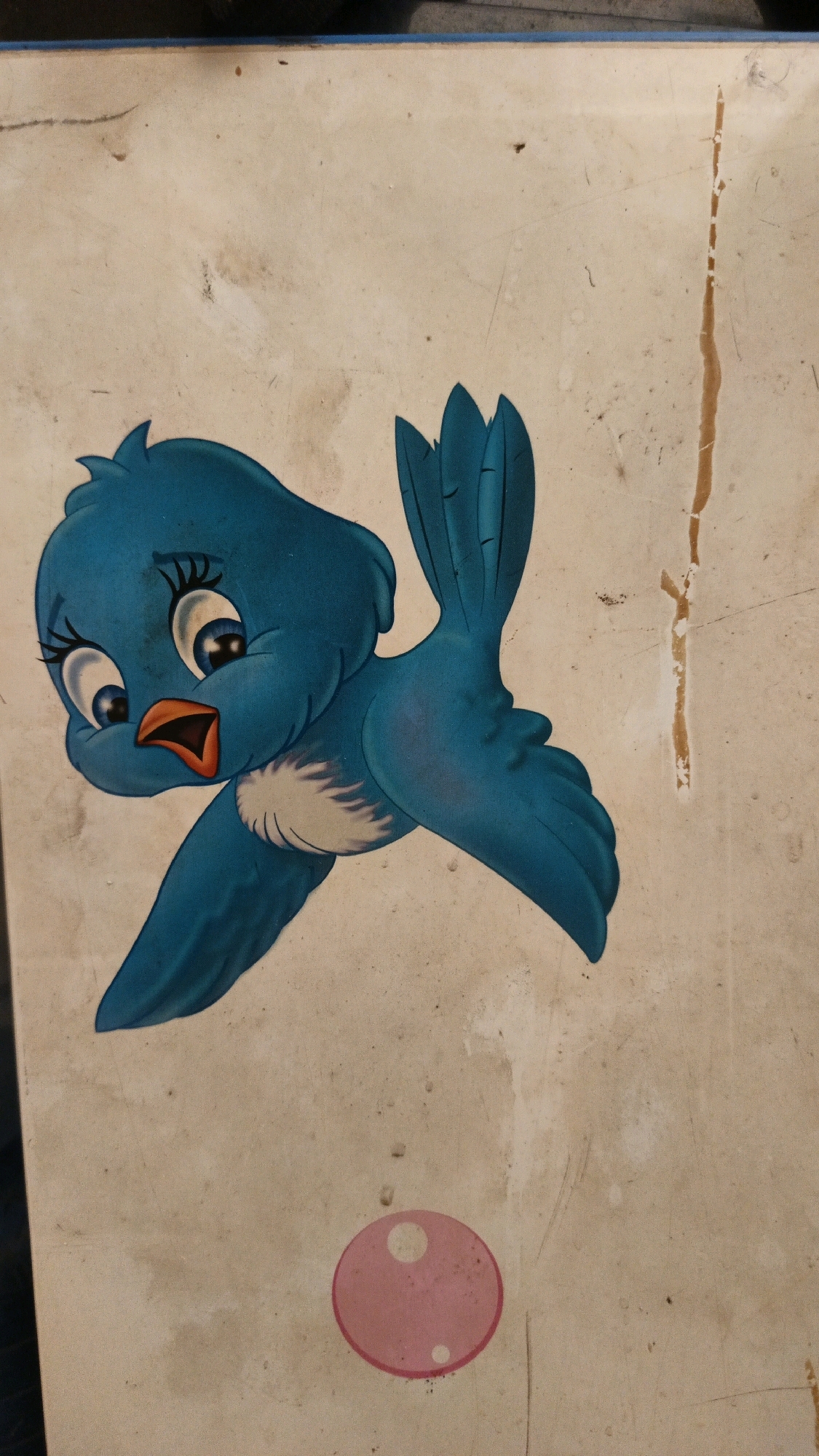 A cartoon illustration of a blue bird is depicted on a light-colored, weathered surface. The bird has large, expressive eyes with long eyelashes, an open orange beak, and white chest feathers. Its wings are spread as if in flight, and its tail feathers are fanned out. Below and to the right of the bird, a pink circle with two smaller white circles inside it is visible, resembling a cartoonish bubble. The background surface shows signs of age and wear, with scuffs, dirt marks, and a vertical brown stain. The image appears to be a sticker or decal on an object. The specific location in North Kuta, Indonesia, cannot be determined from the provided image.
