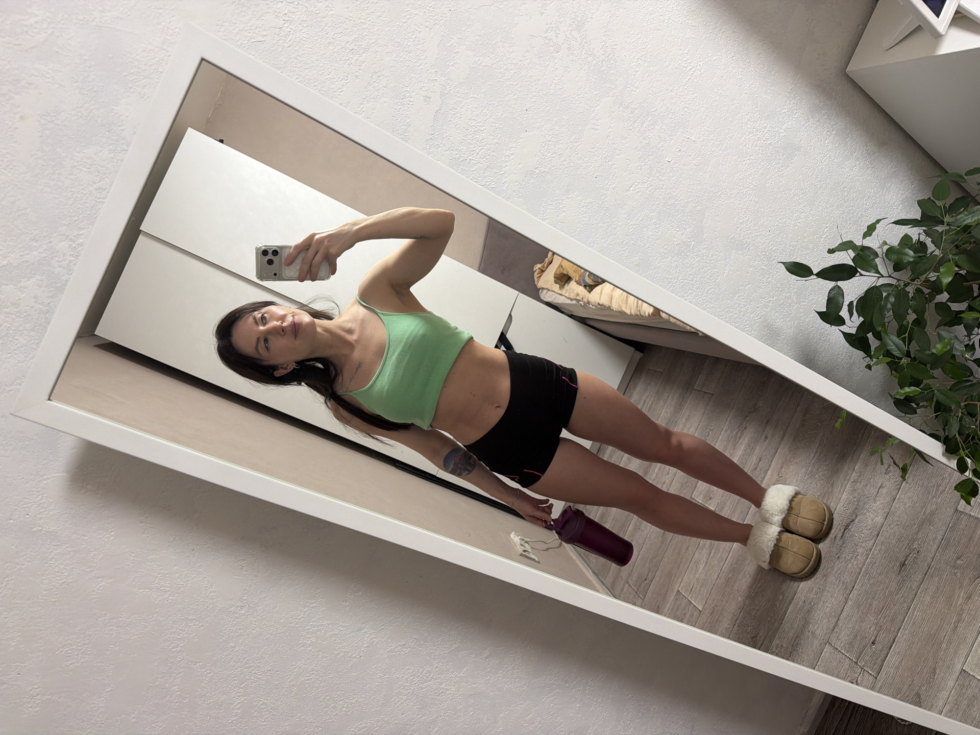 A full-length mirror reflects a woman taking a selfie. She is wearing a light green sports bra and black shorts, along with beige slippers. She's holding a smartphone in her right hand and a purple water bottle in her left. The background includes a white wardrobe, a bed with light bedding, a wooden floor, a wall with a textured surface, and a plant in the corner. The location is suggested to be Perm, Russia, although this is not evident within the image itself.