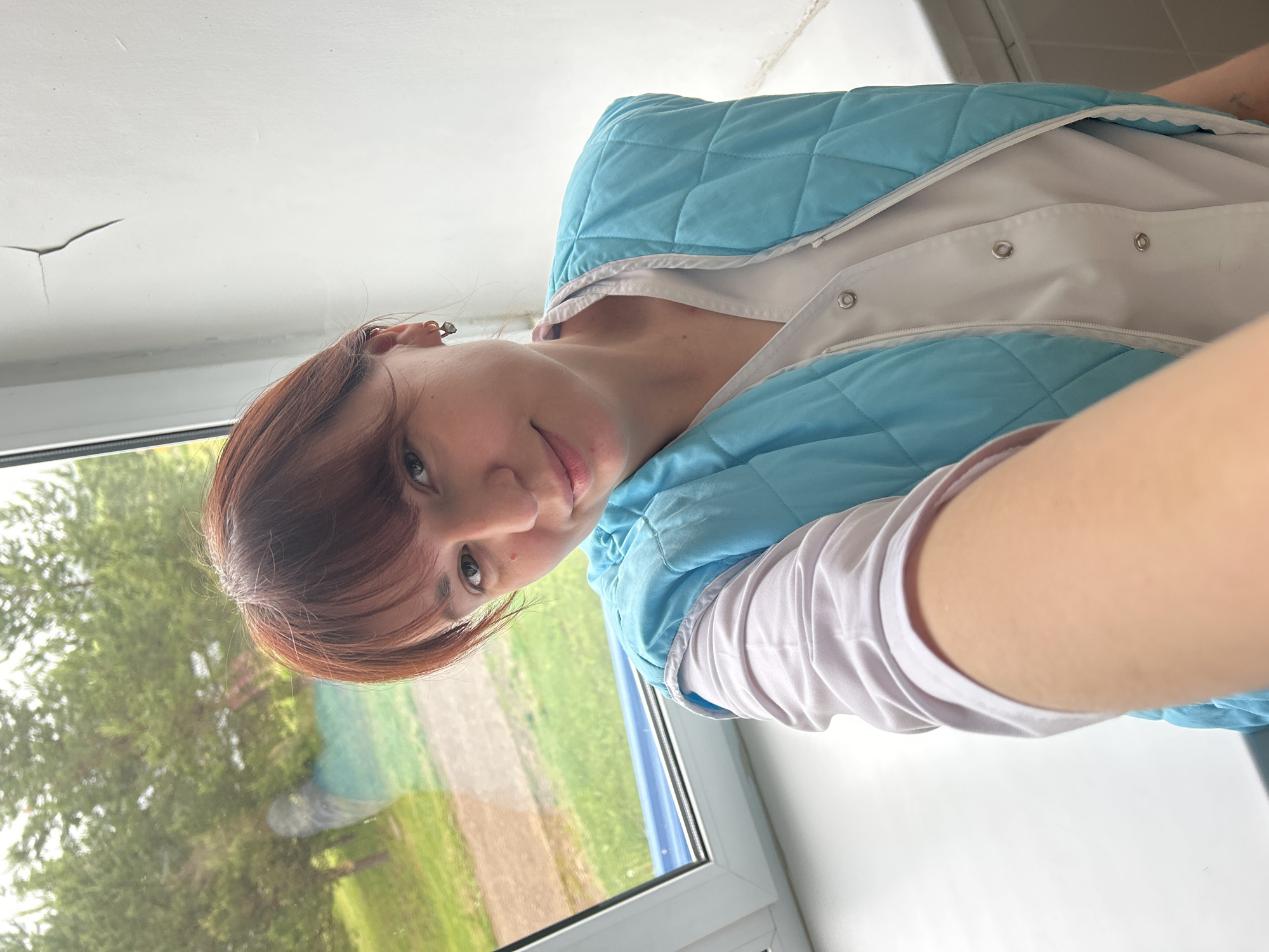 The media file shows a young woman taking a selfie indoors in Фоки, Russia. She has reddish-brown hair with bangs, light-colored eyes, and fair skin, with an earring visible on her left ear. She is wearing a light blue quilted vest over a white long-sleeved top, possibly a uniform. The woman has a calm and slightly smiling expression as she looks towards the camera. She is positioned next to a white-framed window, through which a verdant outdoor scene of green trees and a field is visible, suggesting a rural or natural setting. Above the window, a white ceiling with a noticeable crack is visible. The scene is illuminated by natural daylight, indicating it is daytime. No visible text is present in the image.