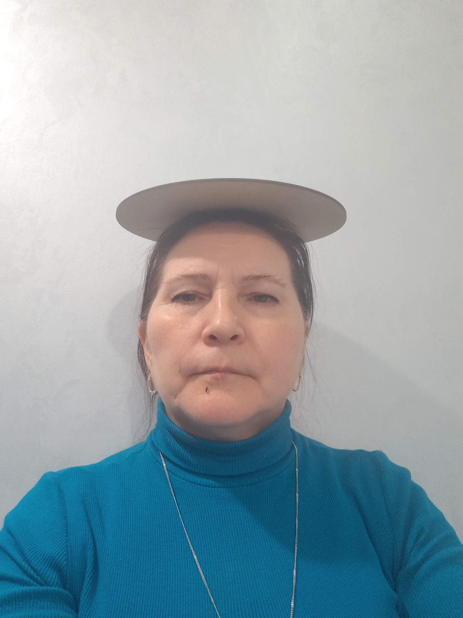 A woman is pictured indoors against a light gray background. She is wearing a blue turtleneck sweater and a silver chain necklace. A flat, round, gray object rests on her head. Her expression is neutral. The image location is specified as Sarapul, Russia.