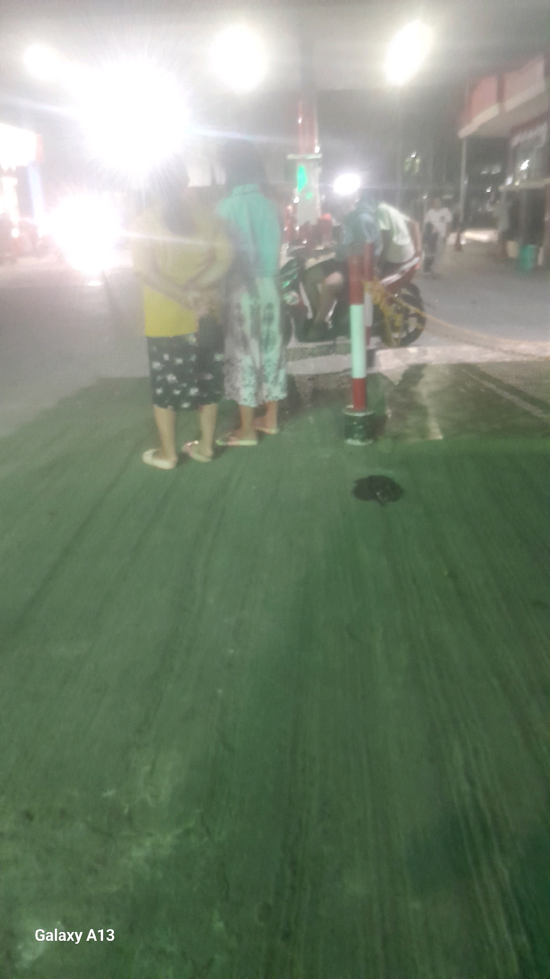 The image depicts a street scene in Jekulo, Indonesia, at what appears to be a gas station. In the foreground, a person is standing, wearing a yellow shirt and patterned shorts, with their legs visible from the knees down. Next to them, another individual is standing, wearing a light blue shirt and patterned pants. Both individuals are facing away from the camera. Further back, a motorcycle is parked, and several other people are visible in the background, though their details are blurred. The environment suggests an outdoor setting, likely a commercial area, with bright lights illuminating the scene from overhead. The ground is paved, and there is a red and white striped pole visible in the mid-ground. The overall image is slightly overexposed due to the bright lights, making fine details difficult to discern. The presence of a gas station sign is implied by the context and lighting.