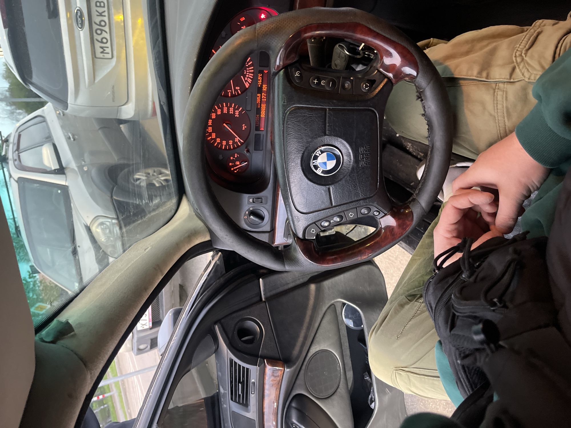 The image depicts the interior of a car, focusing on the dashboard, steering wheel, and a portion of the driver's area. The steering wheel is a BMW model, as indicated by the emblem. The dashboard includes a speedometer, fuel gauge, and other instruments. A Ford is visible through the windshield. The license plate of the Ford reads "M696 KB". The image was taken in the city of Вёшки, Russia. The driver is wearing a green sweatshirt.