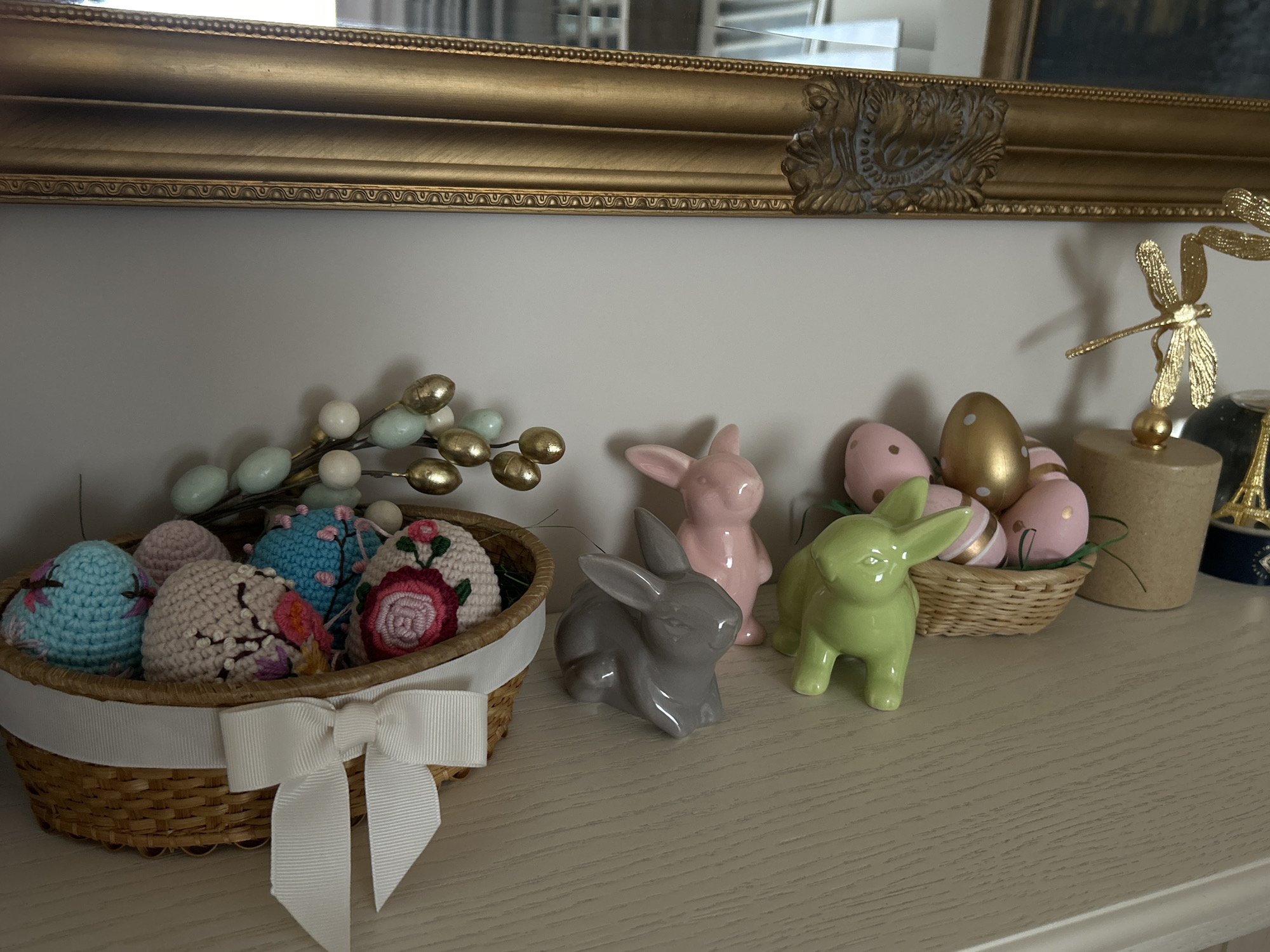 The scene depicts a still life arrangement on a light-colored surface, likely a shelf or mantelpiece, situated indoors. The centerpiece is an Easter-themed collection of objects.
To the left, a woven basket contains several decorated eggs. The eggs are covered in crocheted fabric with floral patterns in pink, blue, and beige colors. A white ribbon is tied around the basket in a bow. Next to the basket is a branch with gold and white decorative berries.
To the right of the basket are three ceramic bunny figurines in gray, pink, and green. Another small woven basket filled with painted Easter eggs is adjacent to the bunnies. The eggs are primarily pink with gold polka dots, except for one gold egg.
Further to the right, there's a cylindrical container and a small gold-colored Eiffel Tower figurine. Above the arrangement is a gold-framed mirror or painting.
The location is impossible to determine solely based on the image. There are no visual indicators to suggest Plovdiv, Bulgaria, or any specific geographical location.