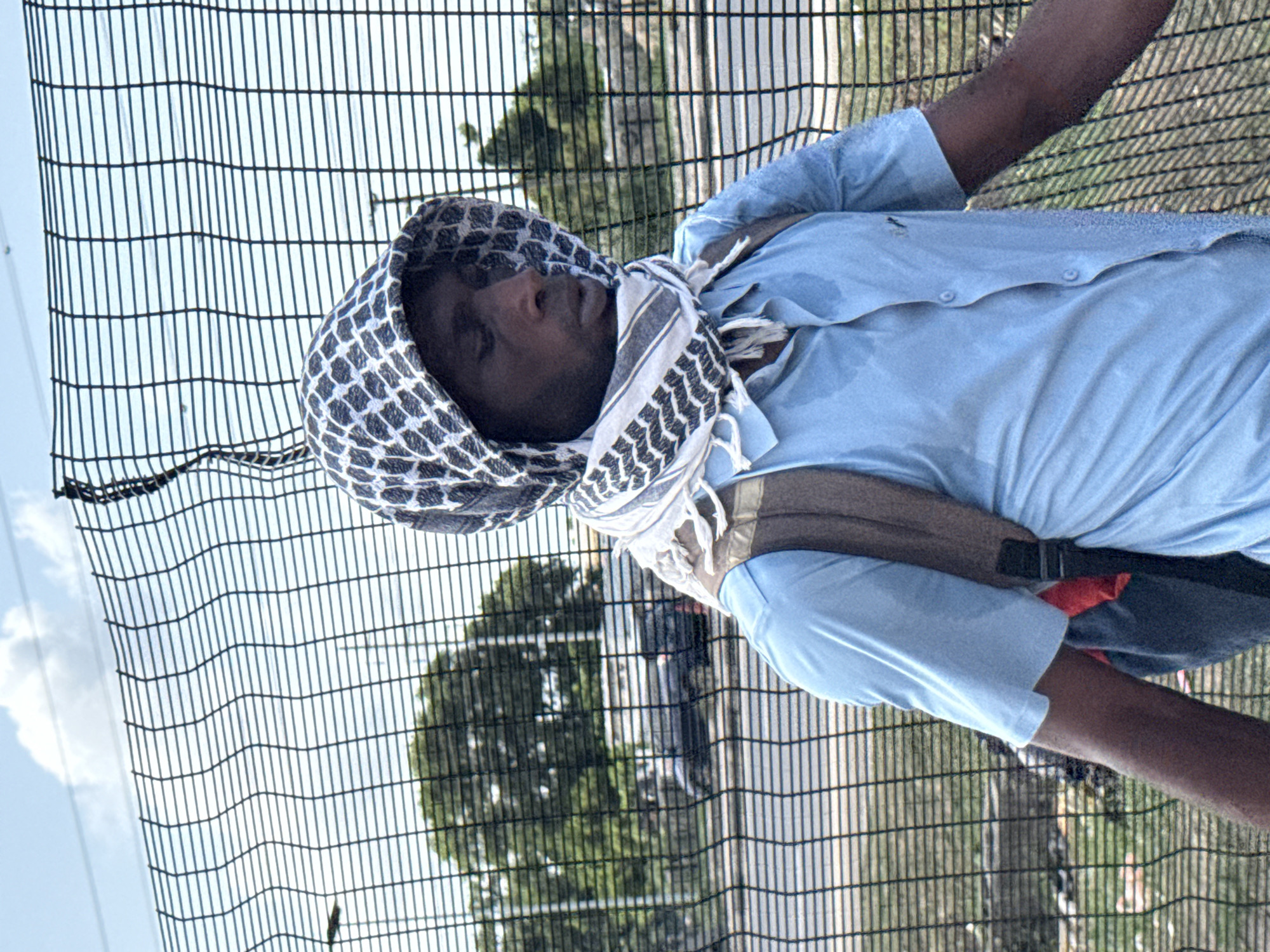 A man is depicted standing outdoors, presumably in Lagos, Nigeria, in a bright daytime setting. He is positioned in front of a dark, vertical mesh fence, which serves as a prominent background element. The man wears a light blue short-sleeved button-up shirt, notable for visible sweat marks that suggest a warm or hot climate. Around his head and neck, he has a black and white patterned keffiyeh-style scarf. A dark backpack is slung over his shoulders, with its straps visible over his shirt. His skin is dark, and his eyes are either partially closed or cast downwards, giving an impression of rest, fatigue, or contemplation.
Through the fence, green trees and distant, blurred elements that could be part of an urban or roadside landscape are visible. The sky, seen through the upper sections of the fence, is bright with some clouds, indicating clear daytime conditions. The fence is made of closely spaced vertical and horizontal dark bars, forming a grid-like pattern. There is no visible text in the image. The overall scene suggests a pause during activity in a warm, outdoor environment.
