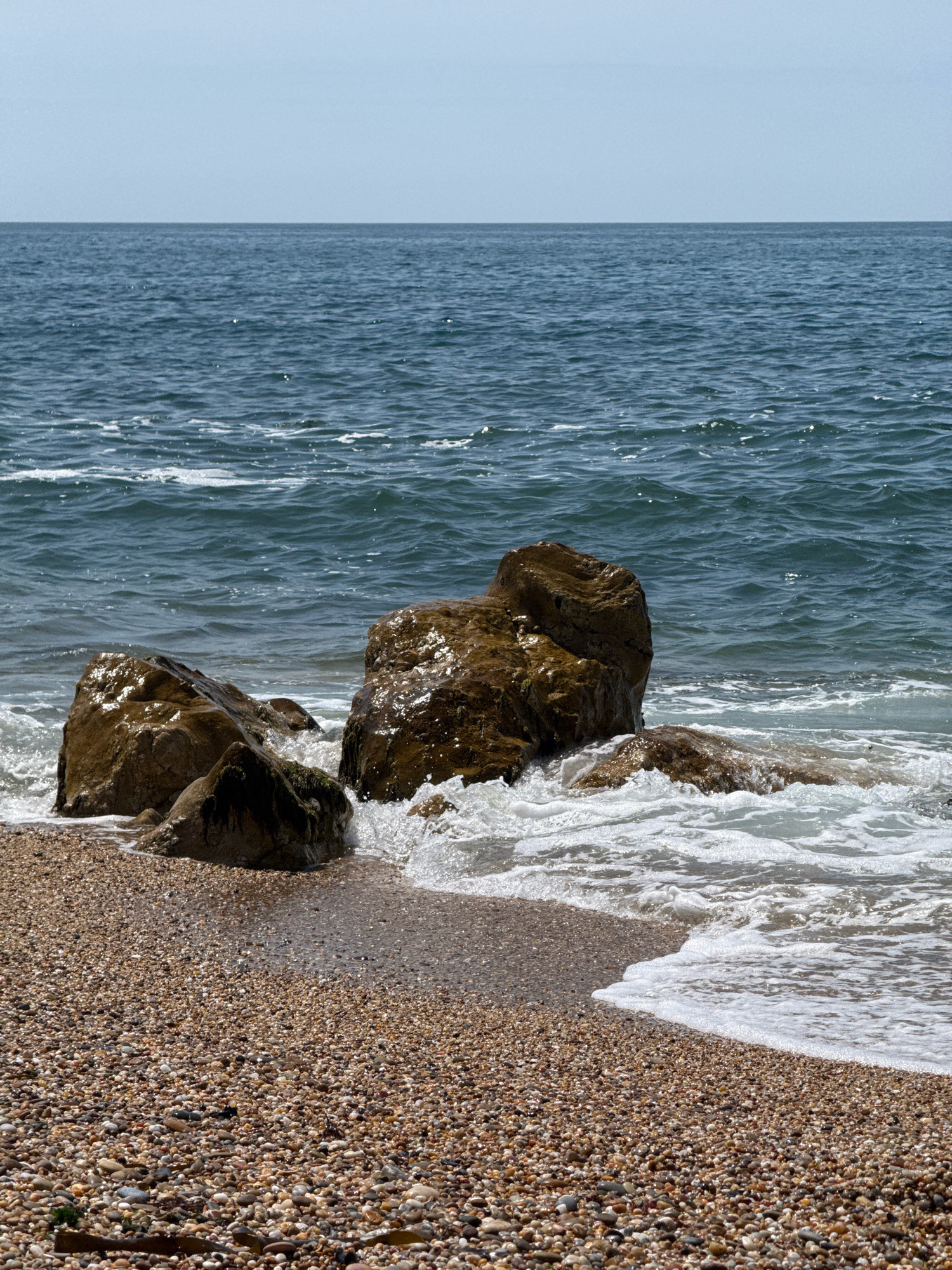 A close-up, eye-level view captures a rocky shoreline meeting the ocean under a clear, pale blue sky. The foreground is dominated by a dense, textured carpet of small, multi-colored pebbles, transitioning into wet sand closer to the water's edge. Several large, wet, brown rocks are partially submerged in the incoming tide, with white foam and spray crashing around them. The ocean itself is a deep blue, with gentle waves and sparkling highlights from the sun. The horizon line is a sharp division between the water and the sky. The scene is set on a beach in Vila Nova de Gaia, Portugal.