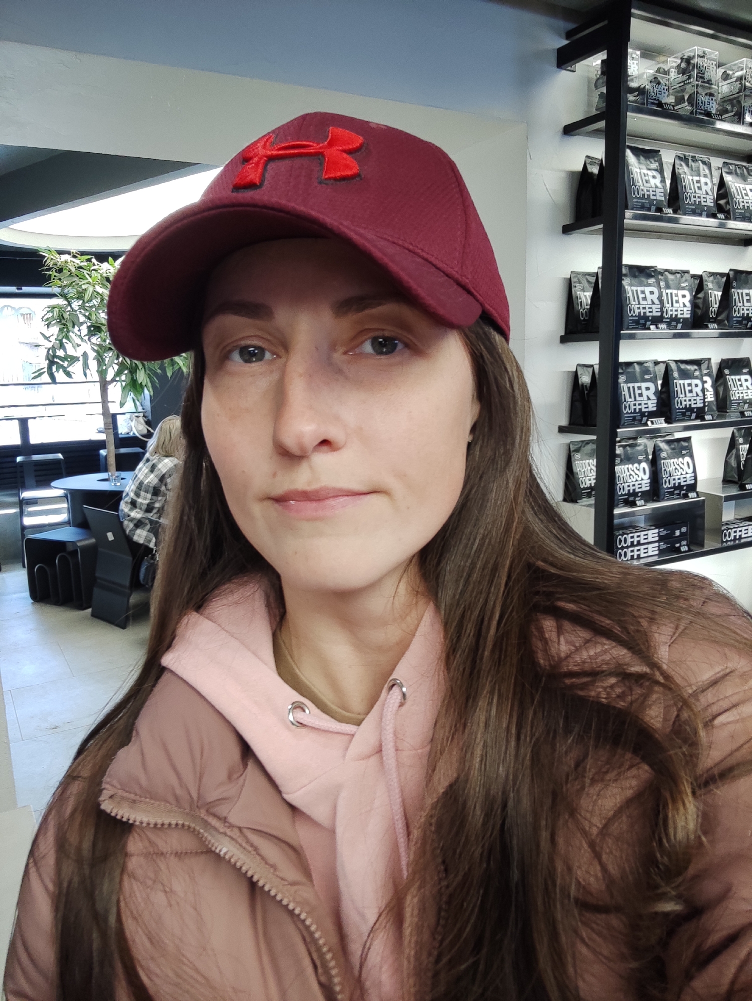 A woman with long brown hair and wearing a maroon Under Armour cap is the primary subject of this selfie-style photograph, taken indoors. She is also wearing a light pink hoodie and a blush-colored puffer jacket. Behind her, on a wall, is a shelving unit filled with packaged coffee bags labeled "Filter Coffee" and "Espresso Coffee". The OCR extracted text indicates the presence of "Coffee" on multiple items. In the background, out of focus, are tables and chairs, with a person seated at one of the tables. The location is a coffee shop, possibly located in Izhevsk, Russia (based on the OCR result mentioning the city name but not visible in the photograph).