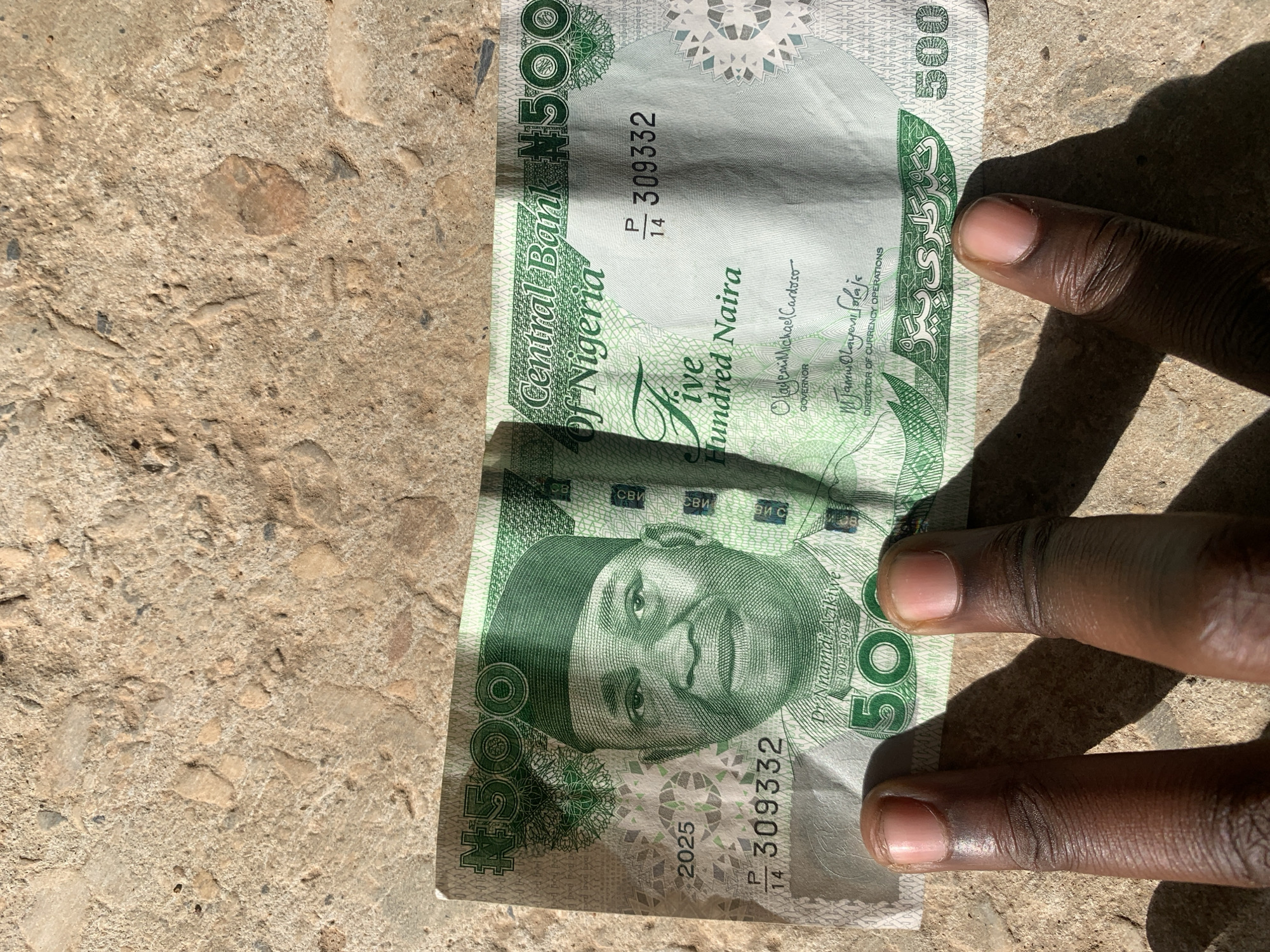 A close-up, eye-level shot captures a Nigerian 500 Naira banknote held by dark-skinned fingers on a textured, light-colored concrete or paved surface. The image is taken outdoors in bright daylight, as evidenced by the strong, distinct shadows cast by the fingers and the banknote. The setting is the New Stadium, Nigeria.
The banknote is predominantly green and white, prominently displaying the portrait of Dr. Nnamdi Azikiwe (1904-1996). The denomination "N500" and "Five Hundred Naira" are clearly visible. Other text on the note includes "Central Bank Of Nigeria", the serial number "P14 309332" (appearing twice), and the signatures of "Olayemi Michael Cardoso GOVERNOR" and "Jamui Olayeni Solaja DIRECTOR OF CURRENCY OPERATIONS". Additional details like "2025" and security markings such as "CB", "CBV", and script in what appears to be Arabic or Ajami are also present. The fingers are holding the note firmly, with the tips of two fingers visible on the left edge and the full length of two fingers on the right edge, slightly obscuring part of the note's design. The ground beneath is rough, composed of varied shades of brown and grey aggregate.