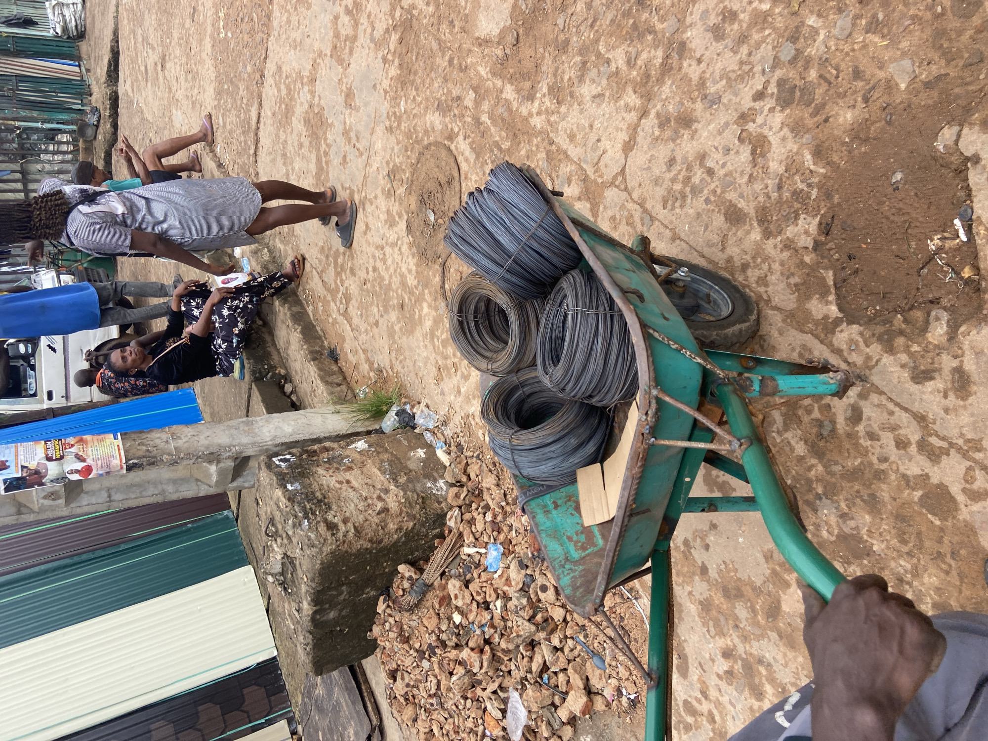 This image depicts a street scene in Ogidi, Nigeria, likely a commercial area, with a focus on a green wheelbarrow filled with coils of wire. In the foreground, a person's hands are visible, gripping the handles of the wheelbarrow, suggesting they are about to move it. The wheelbarrow contains several tightly wound coils of dark, metallic wire. Behind the wheelbarrow, a pile of reddish-brown rubble and rocks is situated next to a concrete structure. Further back, several individuals are present. One woman is seated on a raised step, facing left, wearing a dark top and patterned trousers. Another person, wearing a striped dress, stands to the right, facing away from the camera. In the background, a white truck is partially visible, along with a row of corrugated metal roofing sheets in various colors, leaning against a building or structure. The ground is a rough, earthy surface with patches of mud and scattered debris. A poster with images and text is affixed to a concrete pillar on the left side of the frame. The overall atmosphere appears to be that of an outdoor market or construction site in a bustling urban environment.
