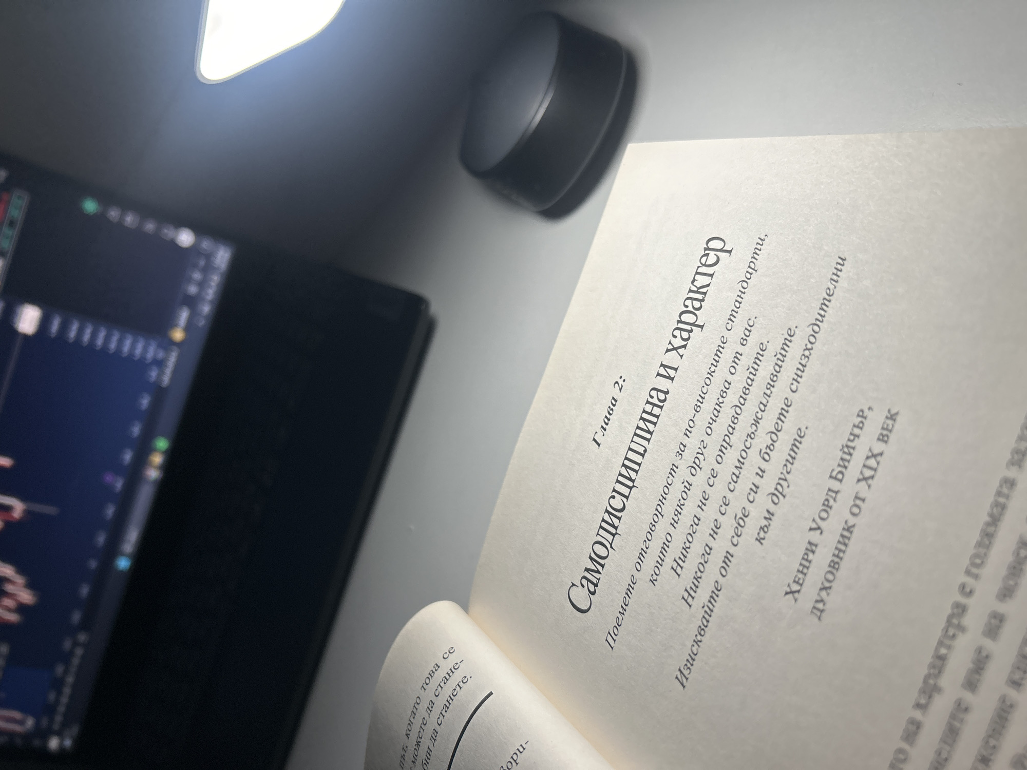 The image is a close-up shot taken indoors, focusing on an open book with text in Cyrillic. The title of the chapter is "Самодисциплина и характер," which translates to "Self-discipline and Character." Beneath the text, the author is identified as Henry Ward Beecher, a spiritual figure from the 19th century. To the left of the book, out of focus, is a computer screen displaying what appears to be a stock market chart. Above the book, there is a black, cylindrical object. The image is illuminated by a bright light source, potentially a lamp, positioned at the top. The background is a light gray surface. The location is not possible to identify from the image.