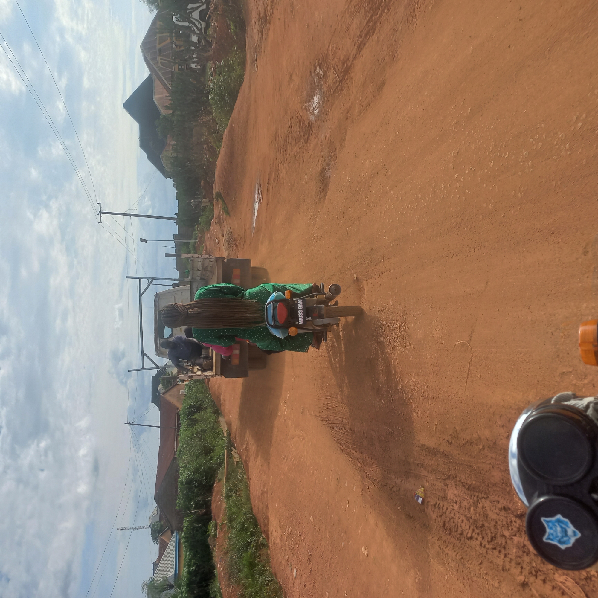 This media file captures a scene on a dusty, reddish-brown dirt road in Mangu Town, Nigeria. The image is oriented sideways, giving a unique perspective. The main subjects are people riding on a motorcycle and a truck.
A person is riding a motorcycle, wearing a vibrant green dress with white polka dots and a blue head covering. Their long, dark hair is visible, cascading down their back. Behind them, another person, wearing a dark blue top, is also seated on the motorcycle. They appear to be traveling together.
In front of the motorcycle is a white truck, also on the dirt road. The truck seems to be carrying some cargo or structure, with metal poles extending upwards. A few other figures are visible in or around the truck. The overall atmosphere suggests everyday transportation or movement within the town.
The setting is rural or semi-rural, with the red earth of the road dominating the foreground. To the sides of the road, there is green vegetation and hints of buildings with reddish roofs, typical of the region. Power lines and poles are also visible against a bright, cloudy sky, indicating daytime. The clouds suggest it might be a partly cloudy day. The presence of tire tracks on the road indicates vehicular movement.
The scene is functional and devoid of overt emotional display. The focus is on the people and vehicles navigating the unpaved road.
Visible text on the back of the motorcycle reads "WUSS GAK".
