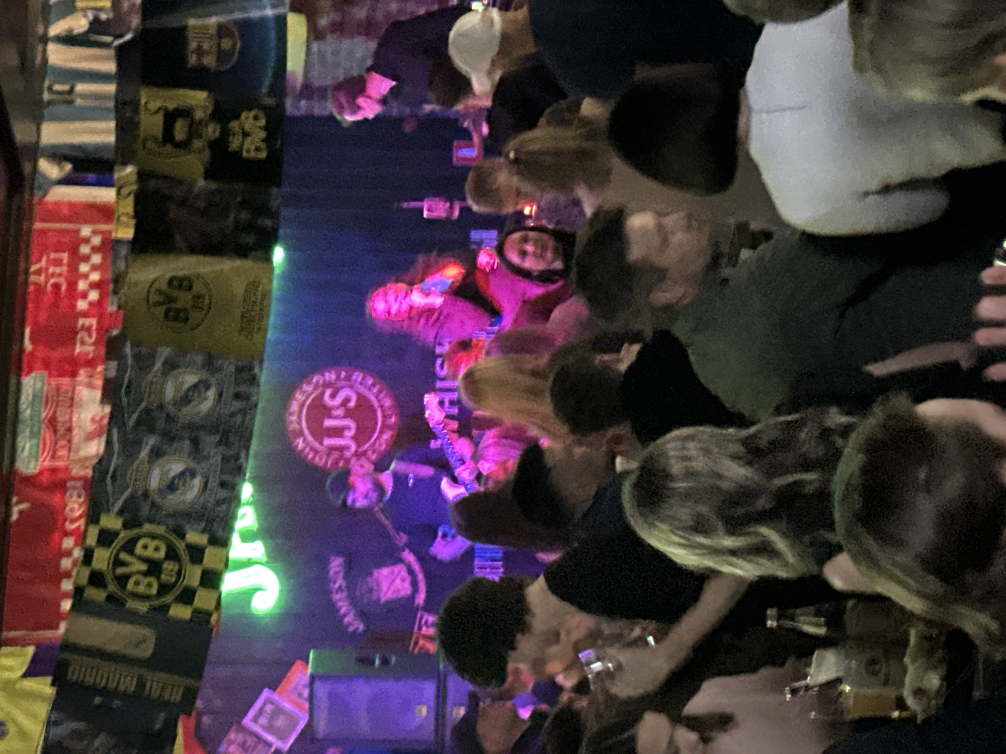 The image shows a crowded interior scene, possibly a bar or a venue with live music. Several people are visible, some of whom appear to be watching a performance on a stage in the background. The stage area is illuminated with neon lights, and the words "JJ's" are visible in a circular logo. The walls and ceiling of the venue are decorated with what appear to be sports scarves and banners, suggesting a sports-themed atmosphere. The people in the foreground are diverse in age and gender, and their interactions are not clearly discernible due to the distance and focus. The lighting is dim, with purple and pink hues dominating the scene, creating a vibrant and energetic ambiance. The location is identified as Minsk, Belarus.