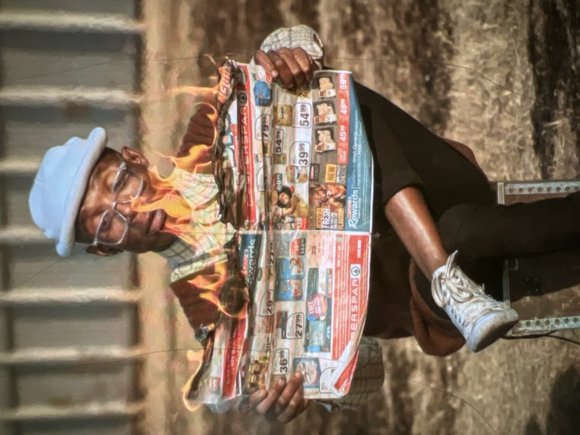 A Black male wearing a light blue bucket hat, glasses, a plaid shirt, and white sneakers sits on a stool, holding a burning newspaper with both hands. Flames engulf the top of the newspaper, obscuring much of its content. The man is looking down at the burning paper with an expression of mild concern or focus. He is dressed in dark trousers. The background appears to be an outdoor environment with dry, brown grass and some indistinct structures, possibly a fence or wall, in the distance. The lighting suggests it is daytime, with the sun casting soft shadows. The image has a slightly grainy, vintage quality, possibly due to the camera or a filter. The scene takes place in Lagos, Nigeria.