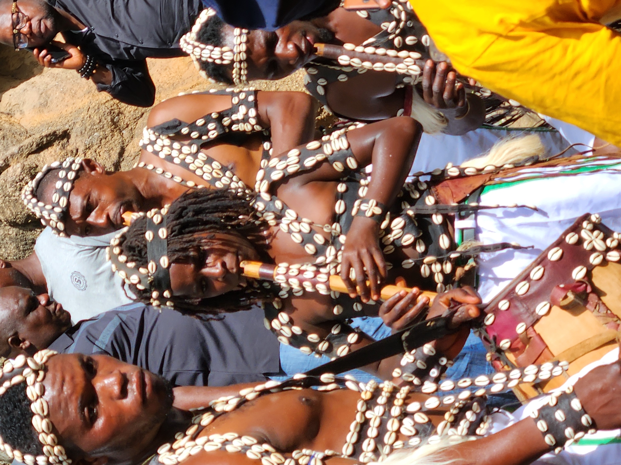 The image shows several people, predominantly dark-skinned, lying on the ground. Most are adorned with white cowrie shells arranged in patterns on their heads, necks, chests, and arms. One person is playing a wooden flute. Some are wearing traditional garments and holding what appears to be a ceremonial drum. One person is holding a mobile phone. The location appears to be outdoors, possibly on a rocky surface.